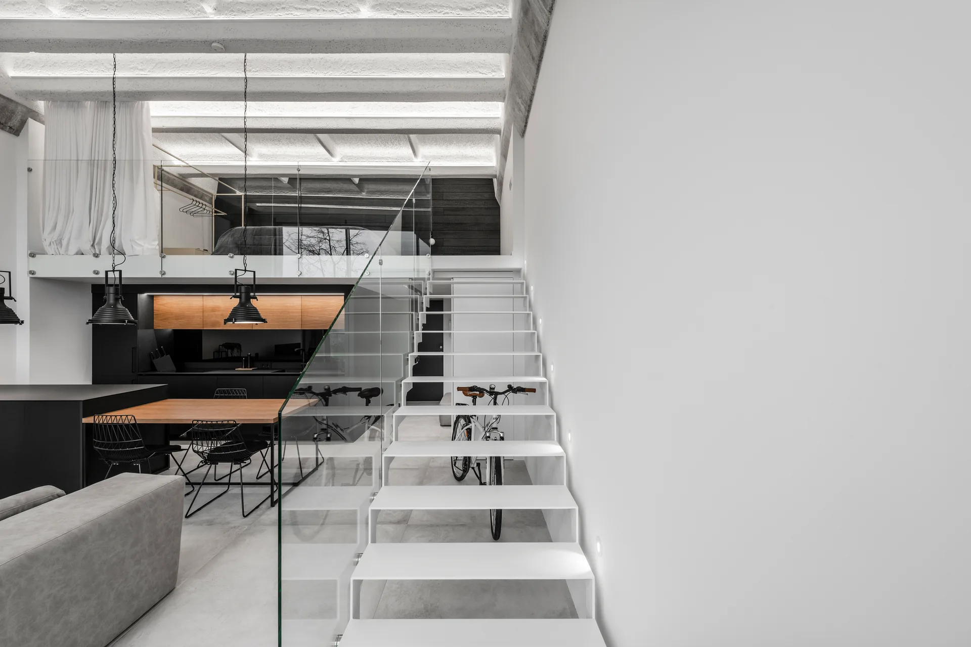 An open-plan loft features a white floating staircase with a glass railing, a wood dining table, and a grey sofa.