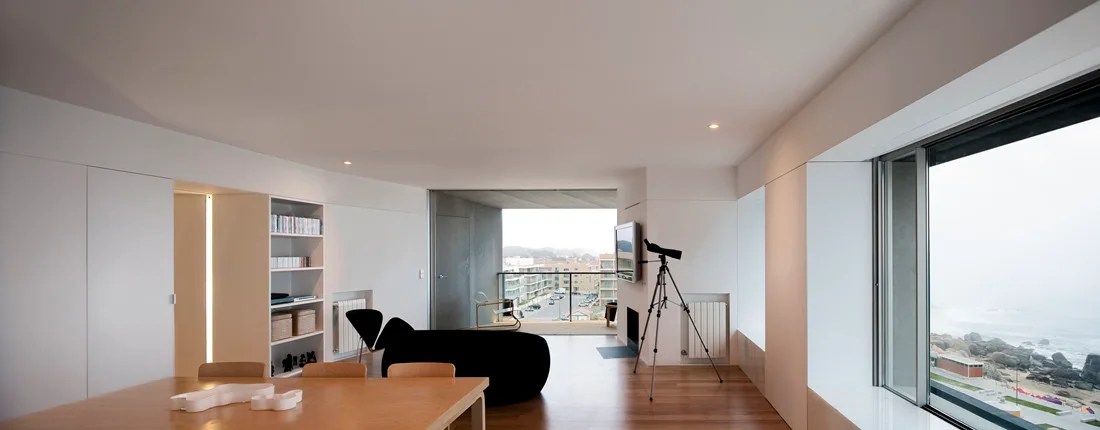 A minimalist living and dining space with white walls, medium-brown wood floors, a curved black sofa, and a large window overlooking the ocean.