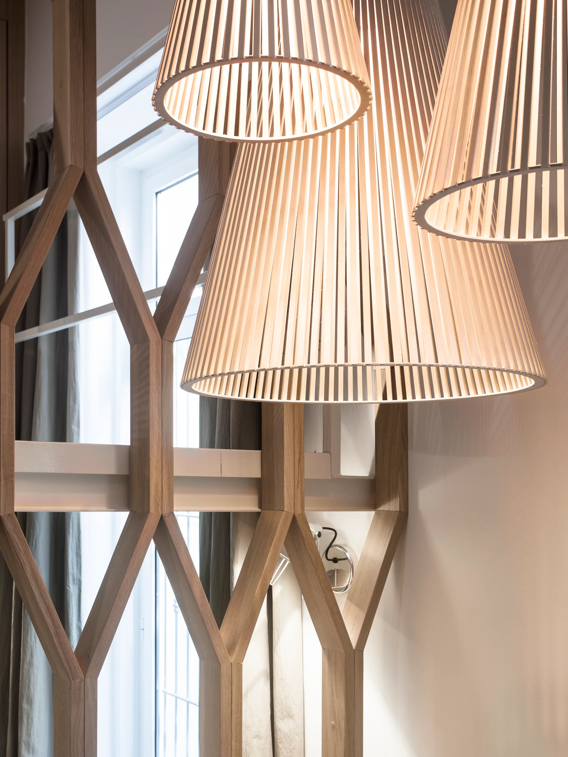 Three illuminated slatted wooden pendant lamps hang in front of an interlocking geometric wood partition screen near a window.