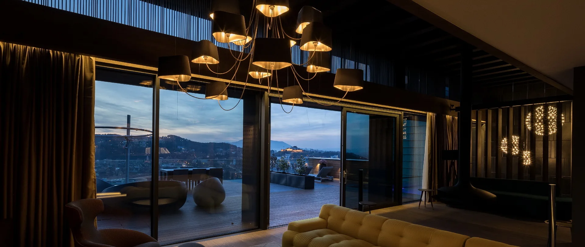 A dimly lit modern living room features a tufted yellow sofa, a large multi-shade chandelier, and sliding glass doors opening to an evening terrace.