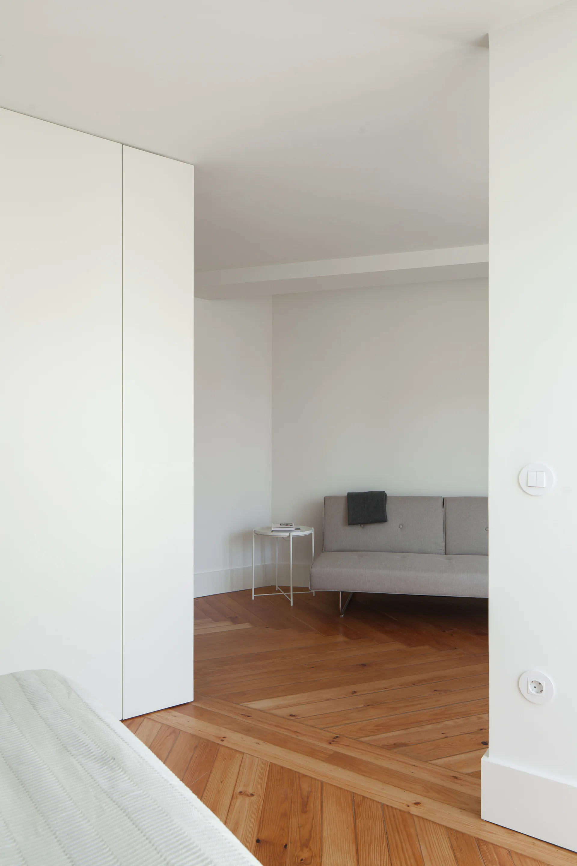 A minimalist bedroom corner features a small grey sofa and a metal side table set on warm herringbone wood floors next to white built-in closets.