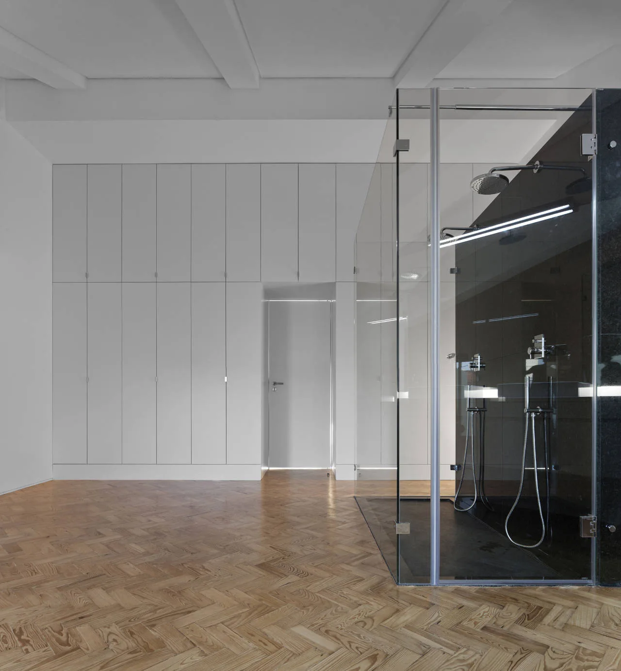 A minimalist bathroom featuring light oak herringbone wood flooring, a glass-enclosed shower with black walls, and a full wall of white storage cabinetry.