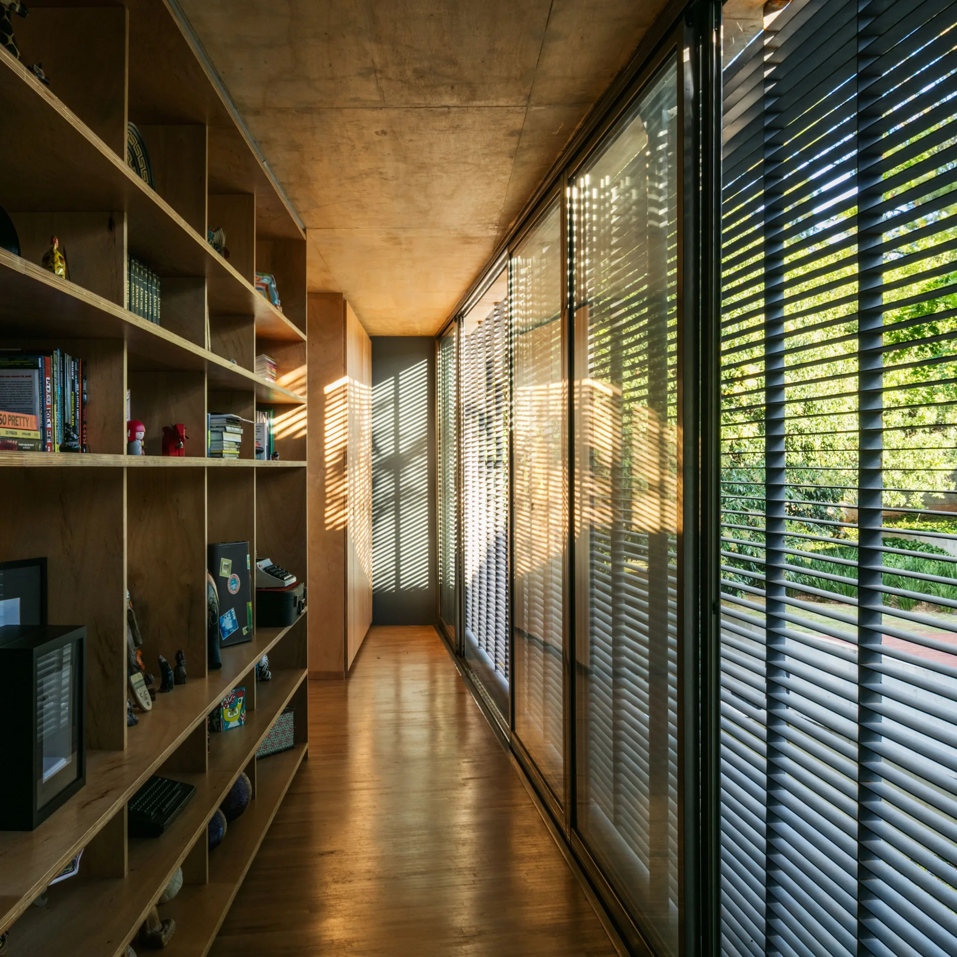 A narrow wood-paneled hallway features a wall of built-in bookshelves and floor-to-ceiling glass doors with horizontal blinds filtering the sunlight.
