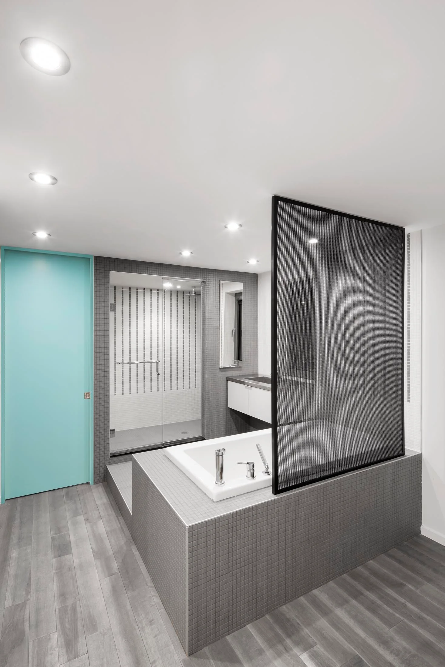 A modern grey tiled bathroom featuring a built-in bathtub with a black mesh privacy screen, a glass shower, and a bright cyan door.