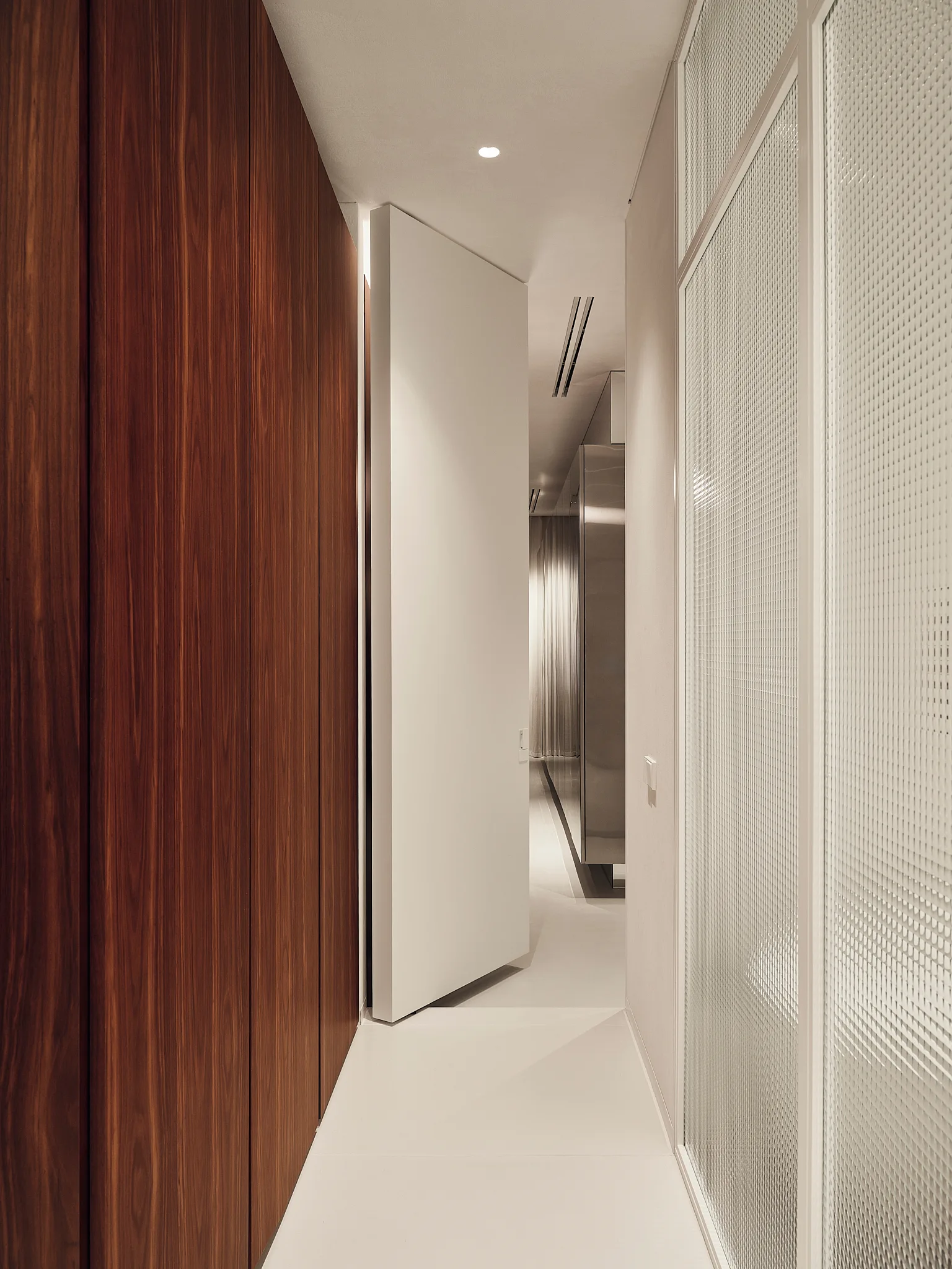 A modern hallway features a large white pivot door flanked by rich vertical wood paneling and a textured grid glass partition wall.