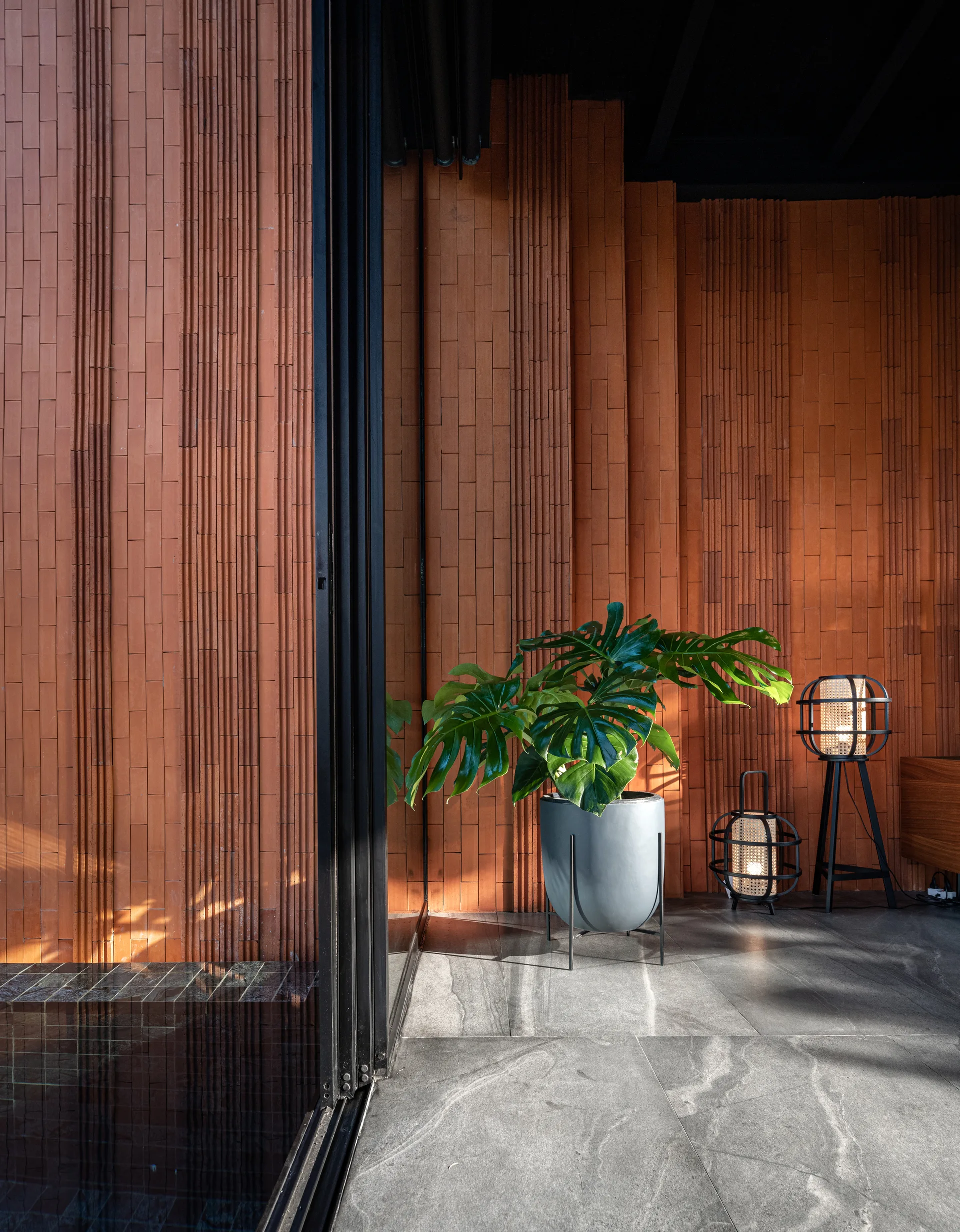 A transition area features a vertically ribbed terracotta brick wall, grey floor tiles, a potted monstera, and black cage-style floor lanterns.