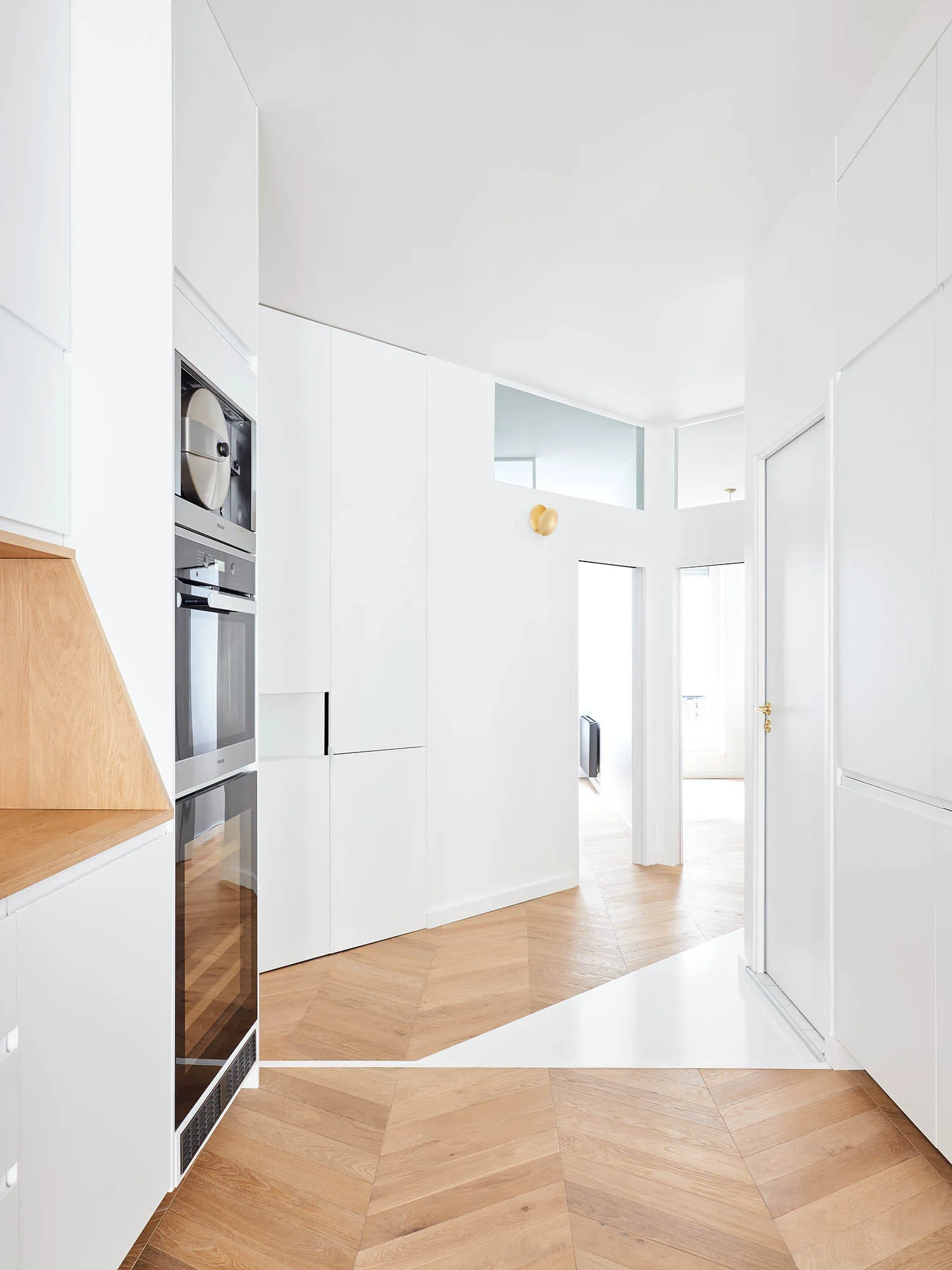 Floor-to-ceiling white minimalist cabinets line a hallway kitchen with integrated appliances, a brass wall sconce, and light wood herringbone flooring.