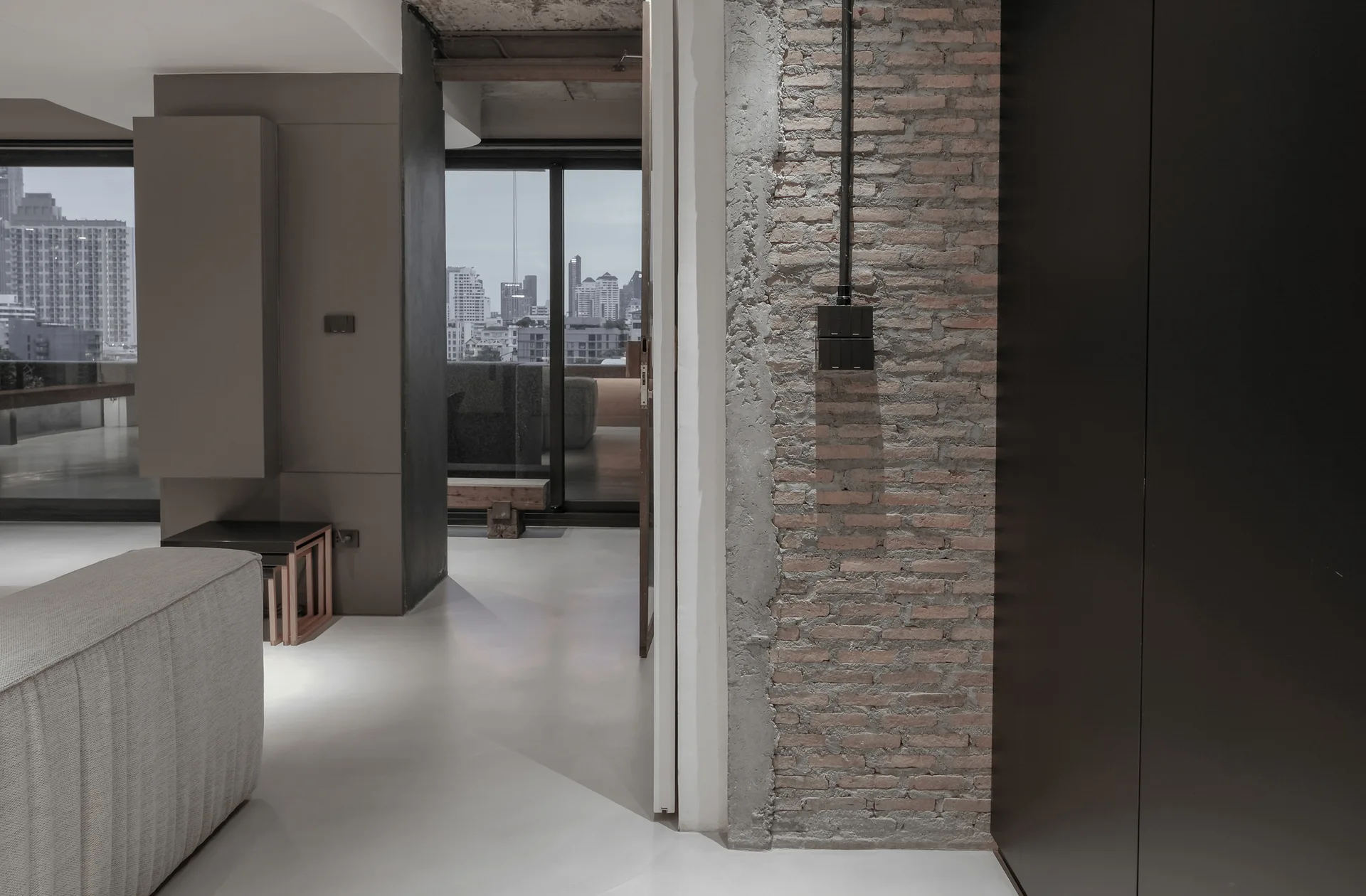 A modern industrial living room features an exposed brick wall, concrete flooring, a grey sofa, and large windows looking out onto a city skyline.