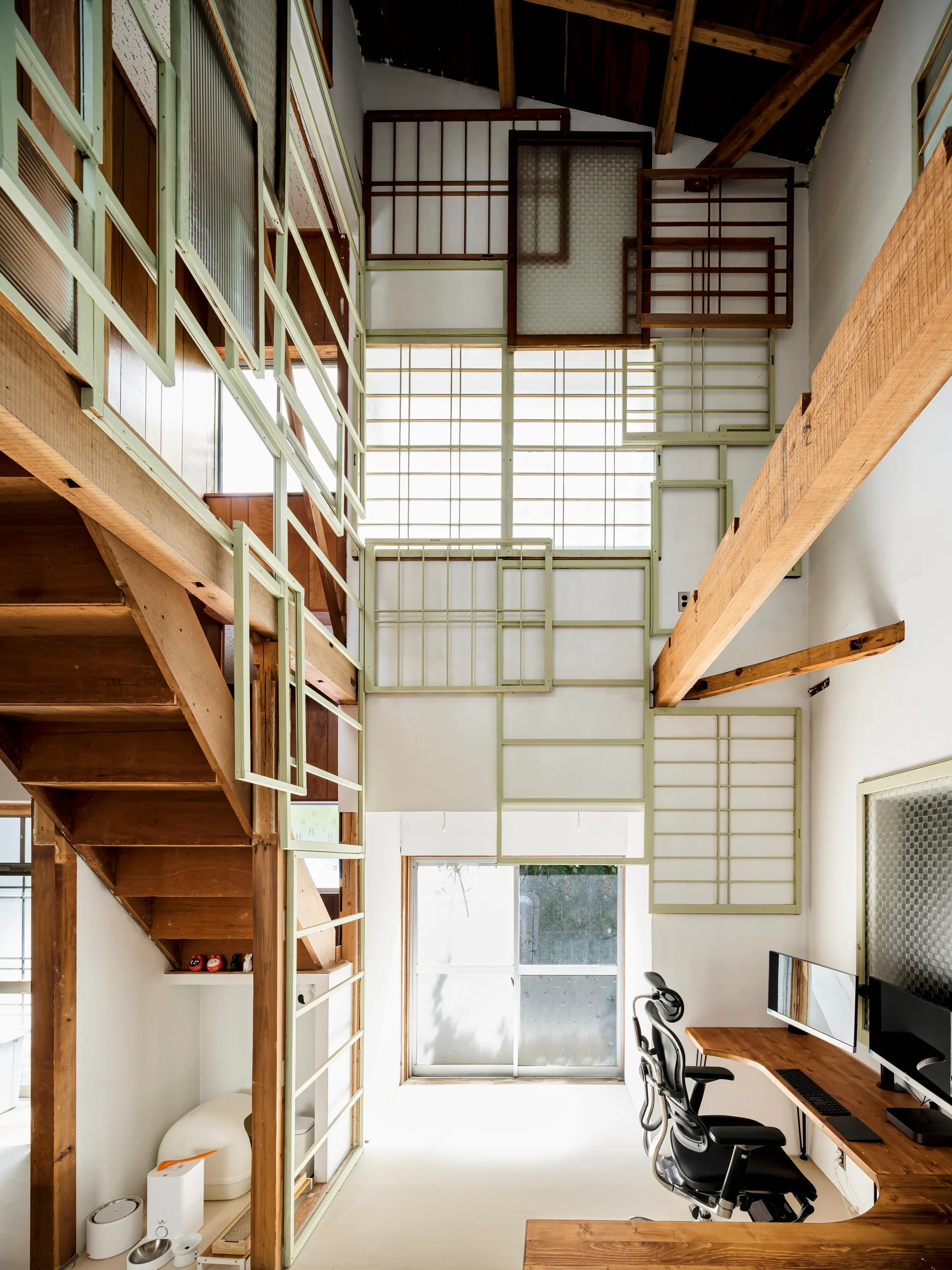 A high-ceiling home office features a curved wooden desk, an open-riser wooden staircase, and a high wall covered in decorative geometric frames.