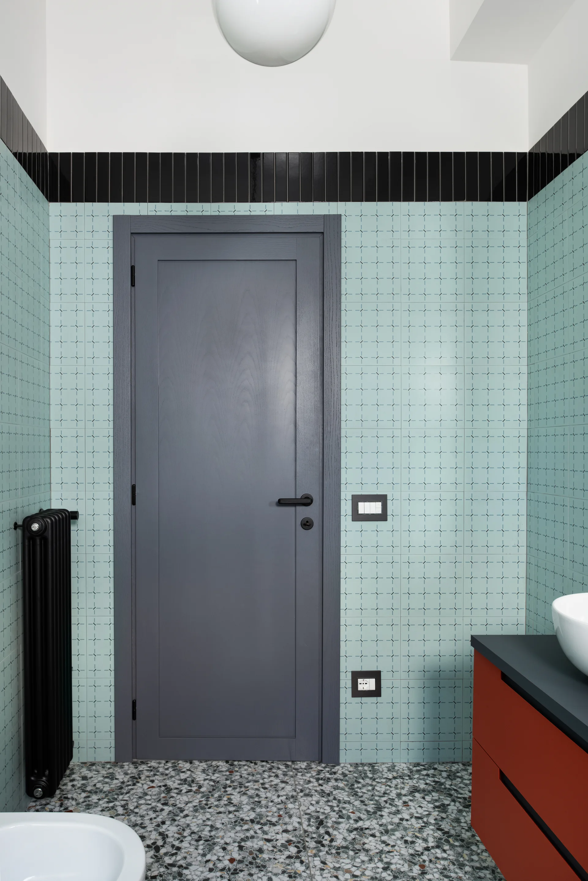 A modern bathroom featuring mint blue tiled walls, a grey door, a red vanity unit, a black radiator, and speckled terrazzo flooring.