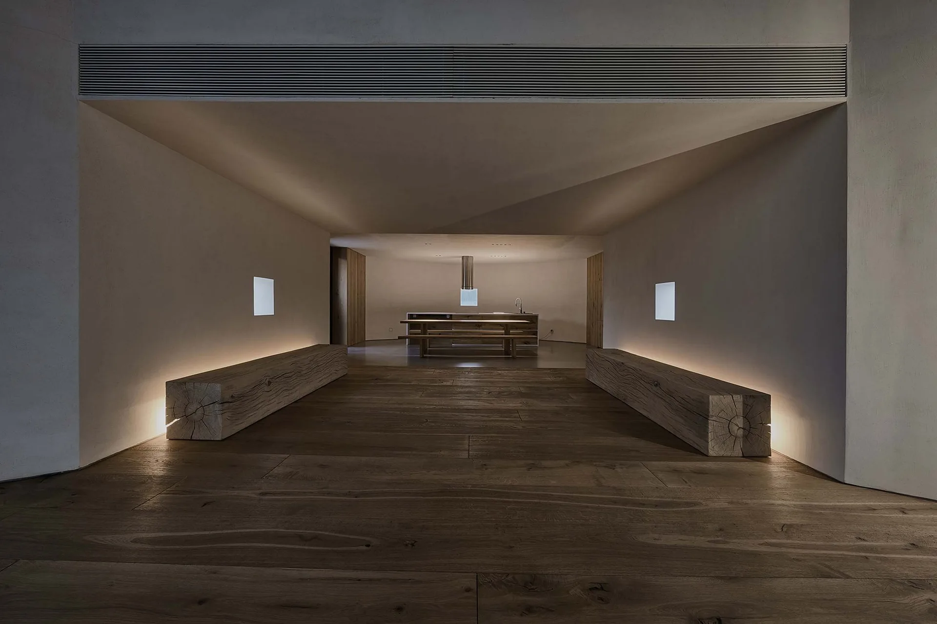 Two massive timber beam benches with warm backlighting line a dark wood corridor leading to a distant kitchen island.