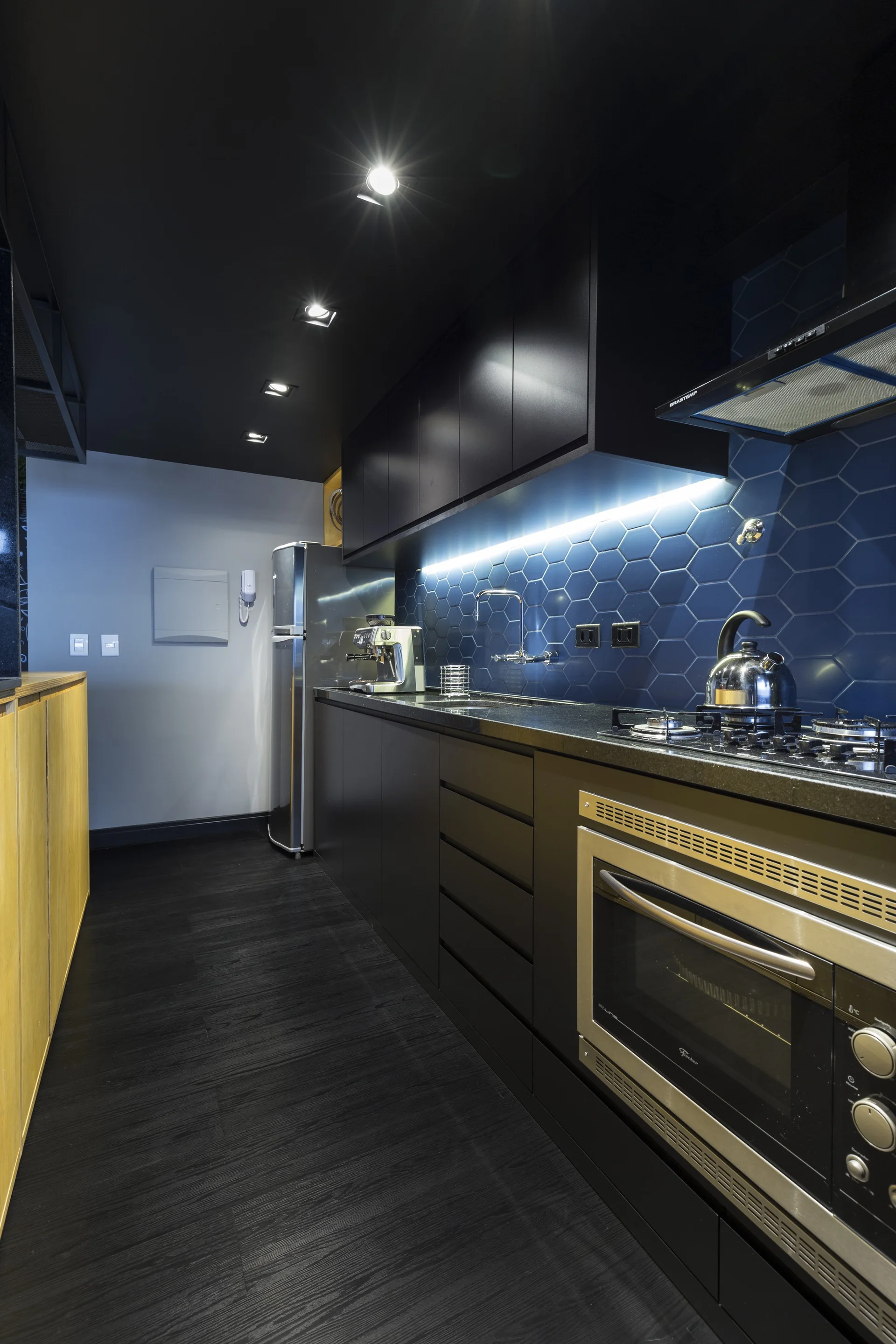 A galley kitchen features matte black cabinets, a black ceiling, and a blue hexagon tile backsplash illuminated by under-cabinet strip lighting.
