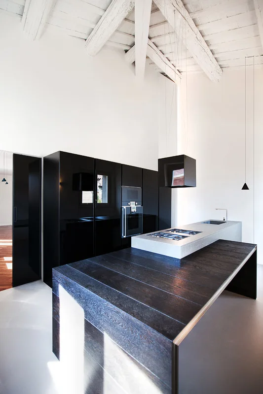 A minimalist kitchen features sleek black cabinetry, a multi-level wood and stone island, and a vaulted ceiling with painted white beams.