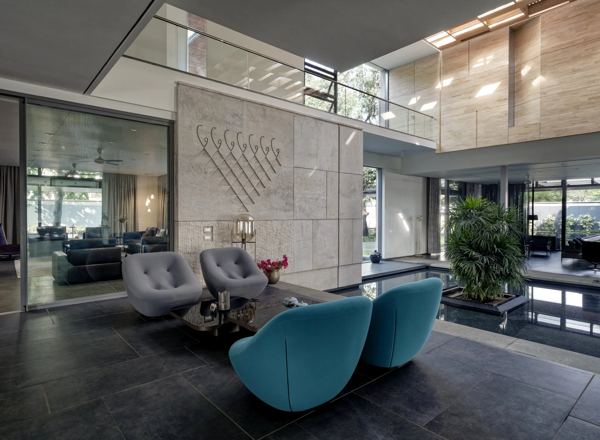 A modern living area with blue and grey lounge chairs around a coffee table, a large stone feature wall, and an indoor pool surrounding a green plant.