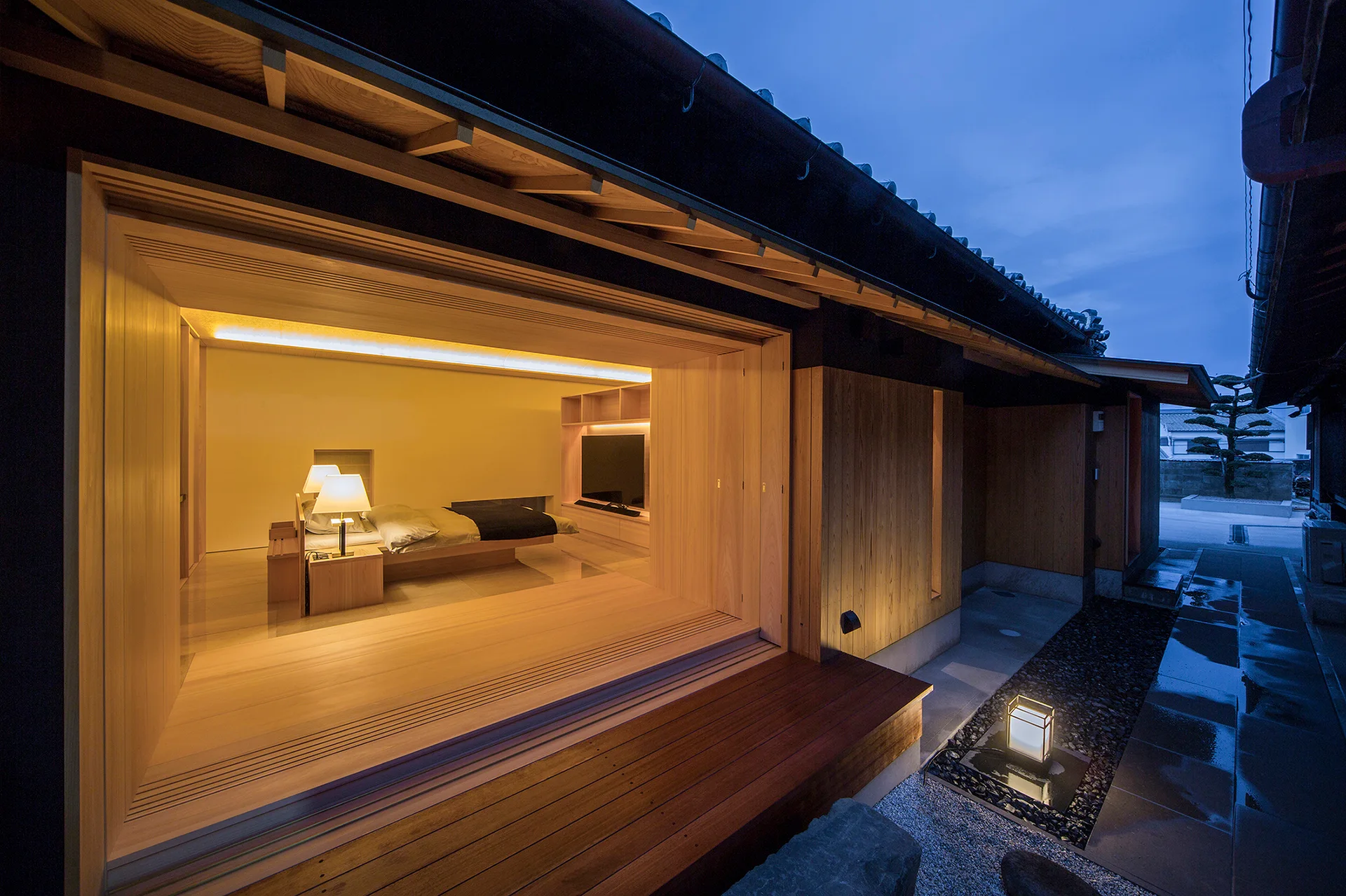 A minimalist wood-paneled bedroom features a low platform bed and opens fully to an exterior wooden deck and gravel path at dusk.