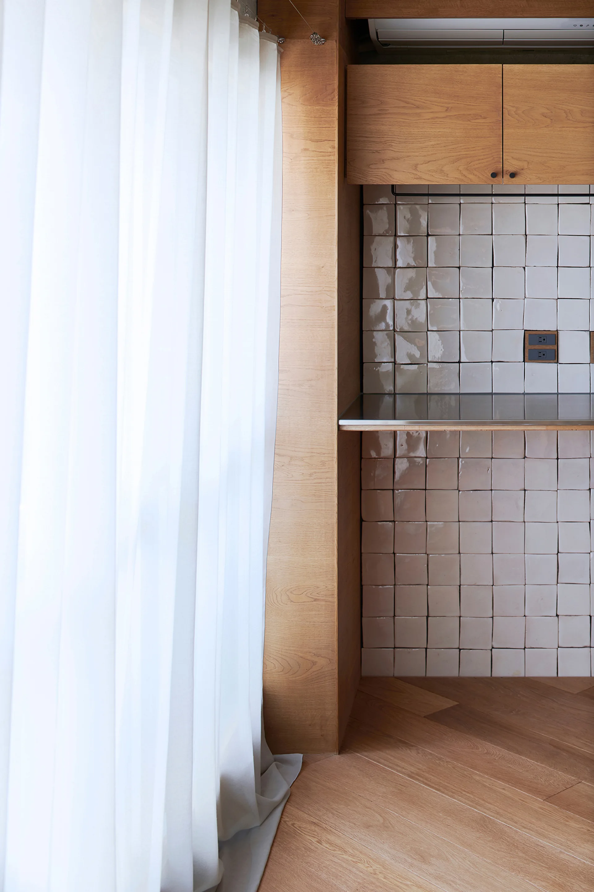 A kitchen corner featuring light wood cabinets, a glazed square tile backsplash, and herringbone wood floors next to a sheer white window curtain.