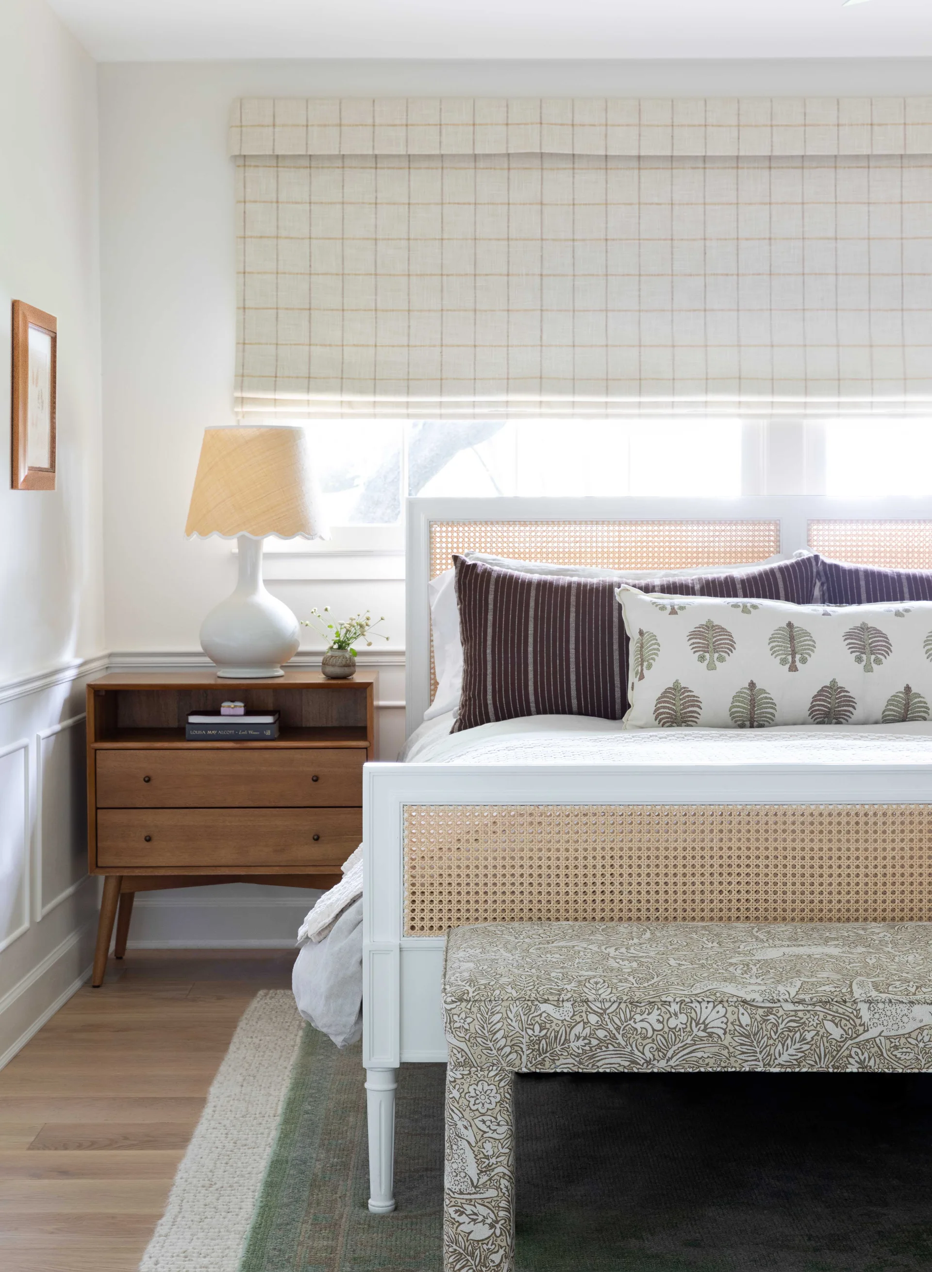 A bedroom featuring a white wooden bed frame with cane panels, a mid-century wooden nightstand, and a checkered Roman shade over the window.