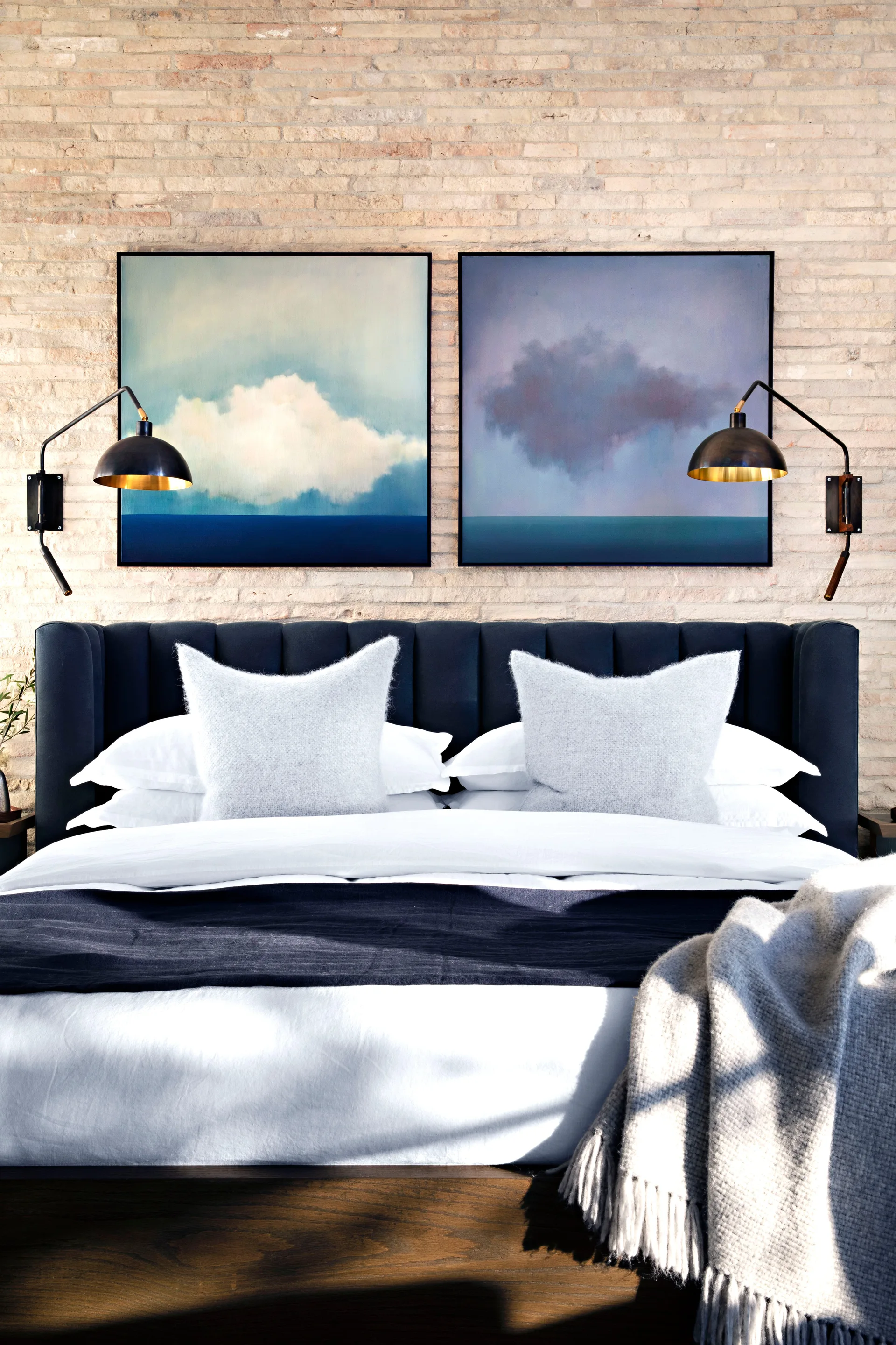 A bedroom features a navy channel-tufted headboard against an exposed brick wall, accented by a matching pair of square art pieces and black sconces.