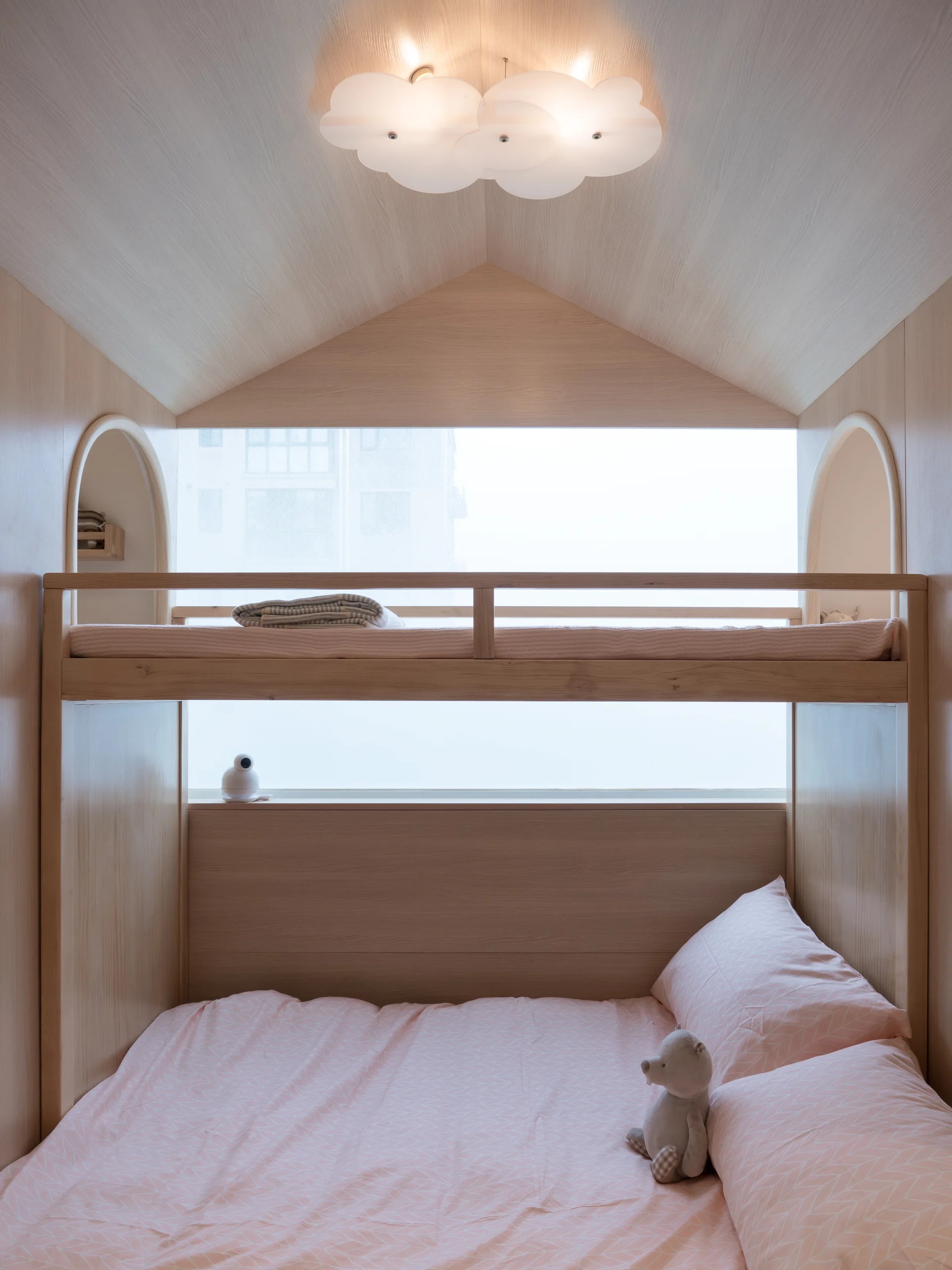 A children's room features a built-in light wood bunk bed set against a window, with pink geometric bedding and a cloud-shaped ceiling light.