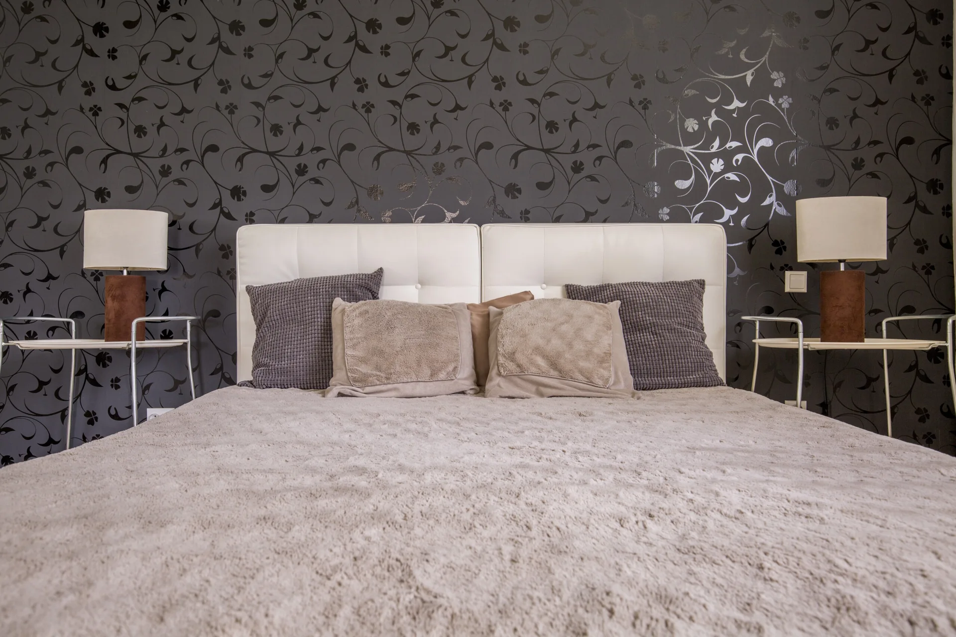 A bedroom features a white tufted headboard on a bed with brown pillows set against dark grey floral wallpaper with two bedside lamps.