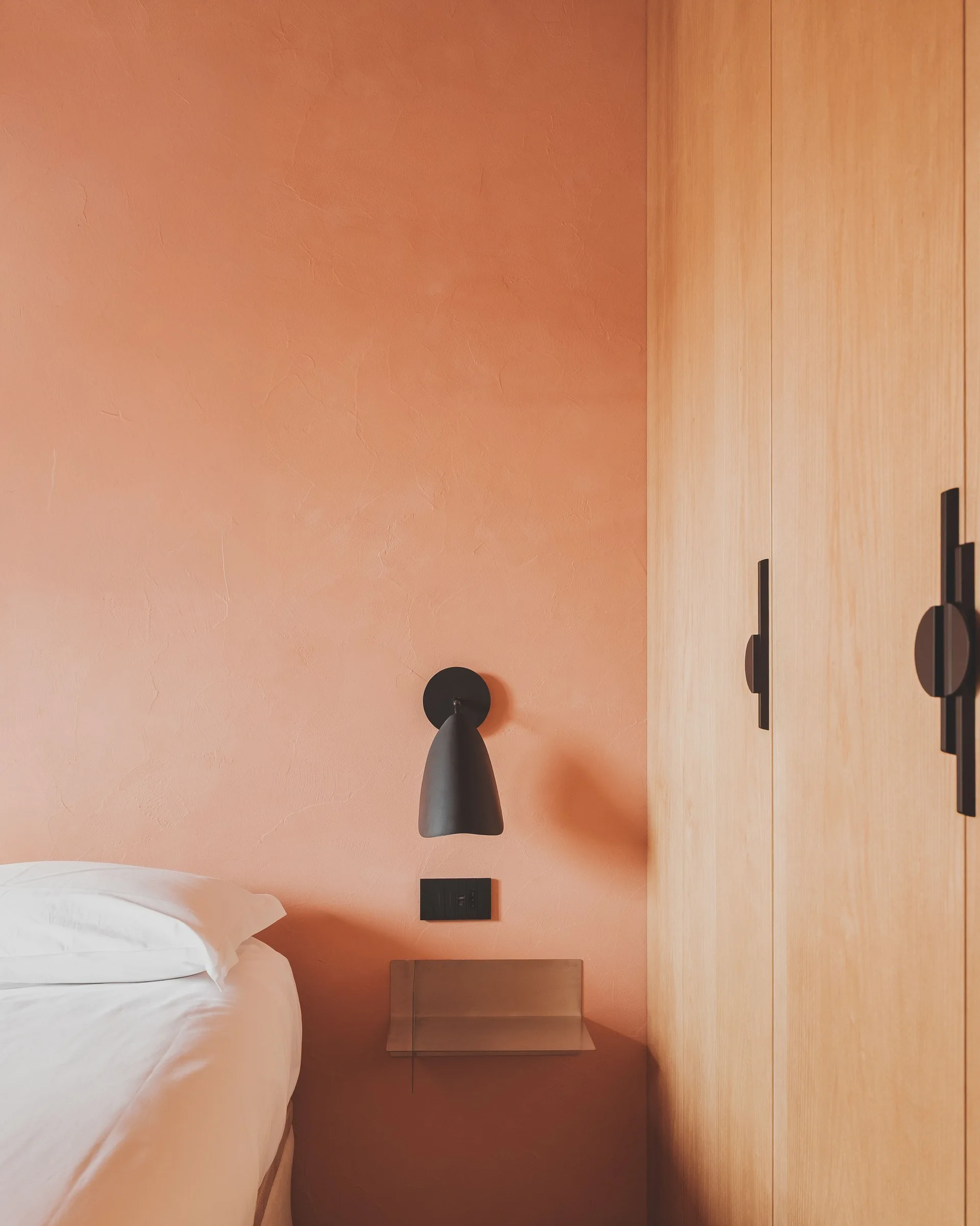 A minimalist bedroom corner featuring a textured peach plaster wall, a black wall sconce, and a light wood wardrobe with black handles.