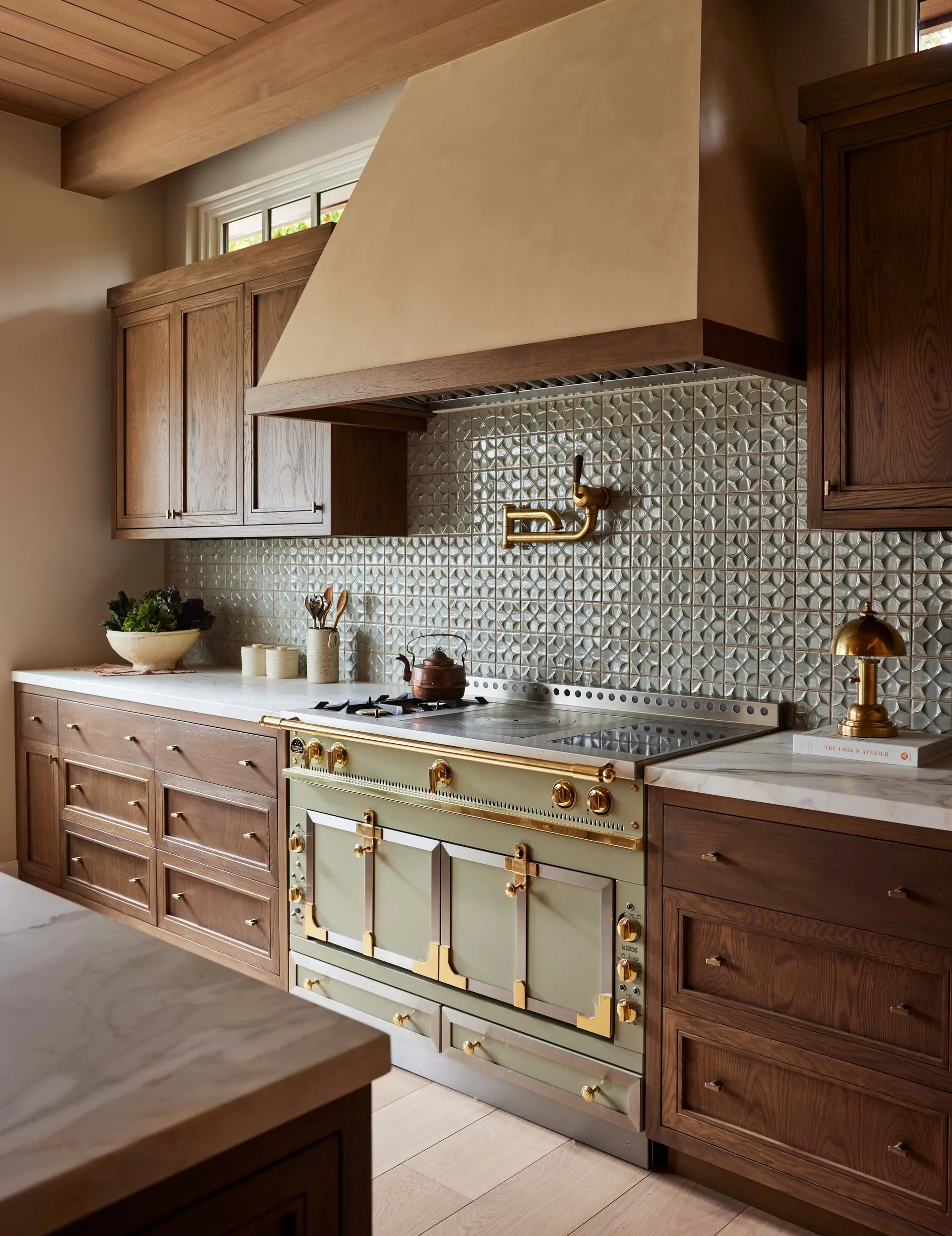 A kitchen featuring a sage green range, wood cabinets, a geometric tiled backsplash, and a large plaster hood under a beamed ceiling.