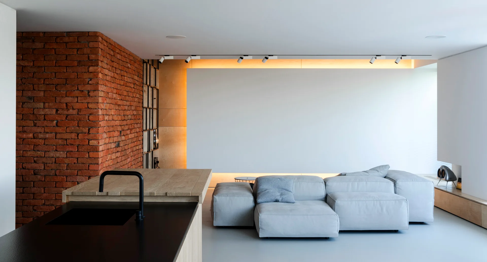 A modern open-plan living area features a modular grey sofa, a large red brick accent wall, and a glowing backlit wall panel behind the seating.