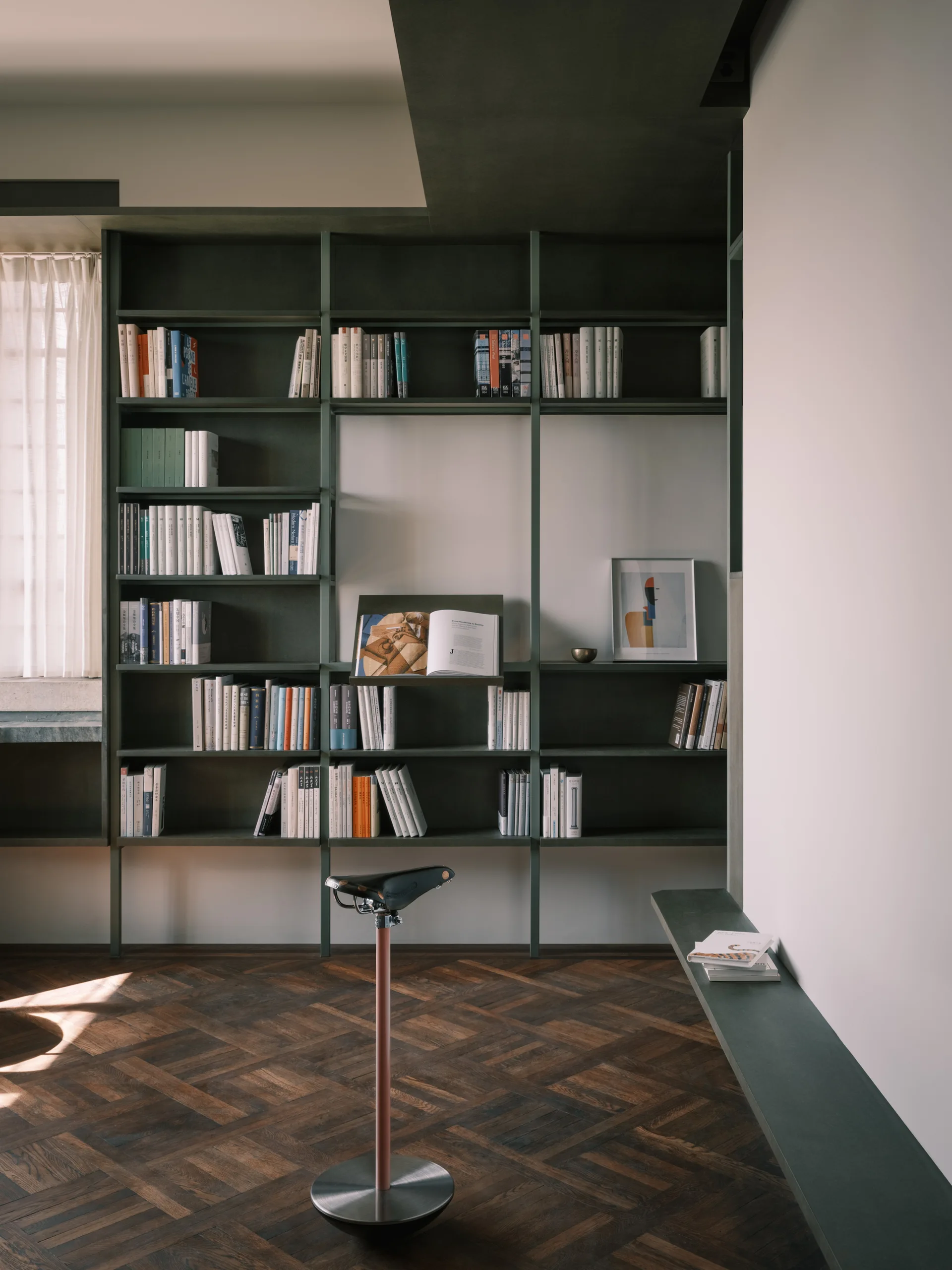 A minimalist study features a dark green floor-to-ceiling shelving unit filled with books, a bicycle-seat stool, and dark parquet wood floors.