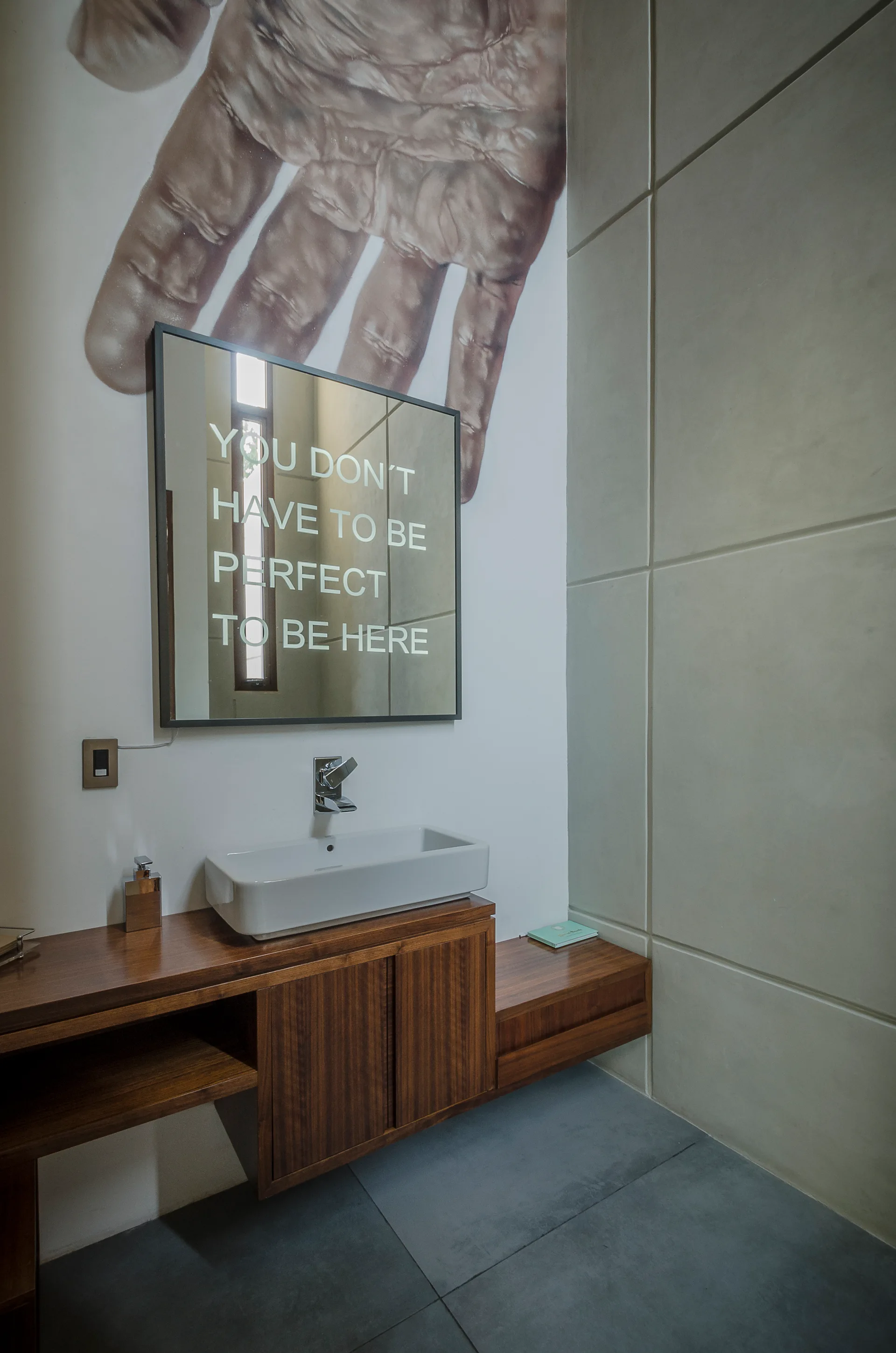 A modern powder room features a floating wooden vanity, a white rectangular vessel sink, concrete-panel walls, and a mirror printed with text.