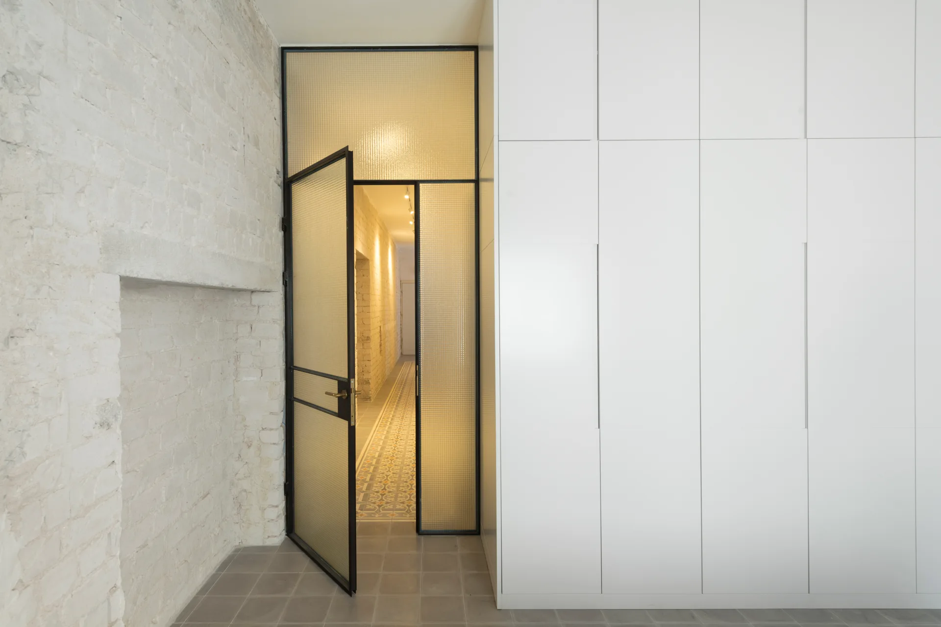 A black-framed textured glass door stands slightly open between a painted white brick wall and a bank of seamless white floor-to-ceiling cabinets.