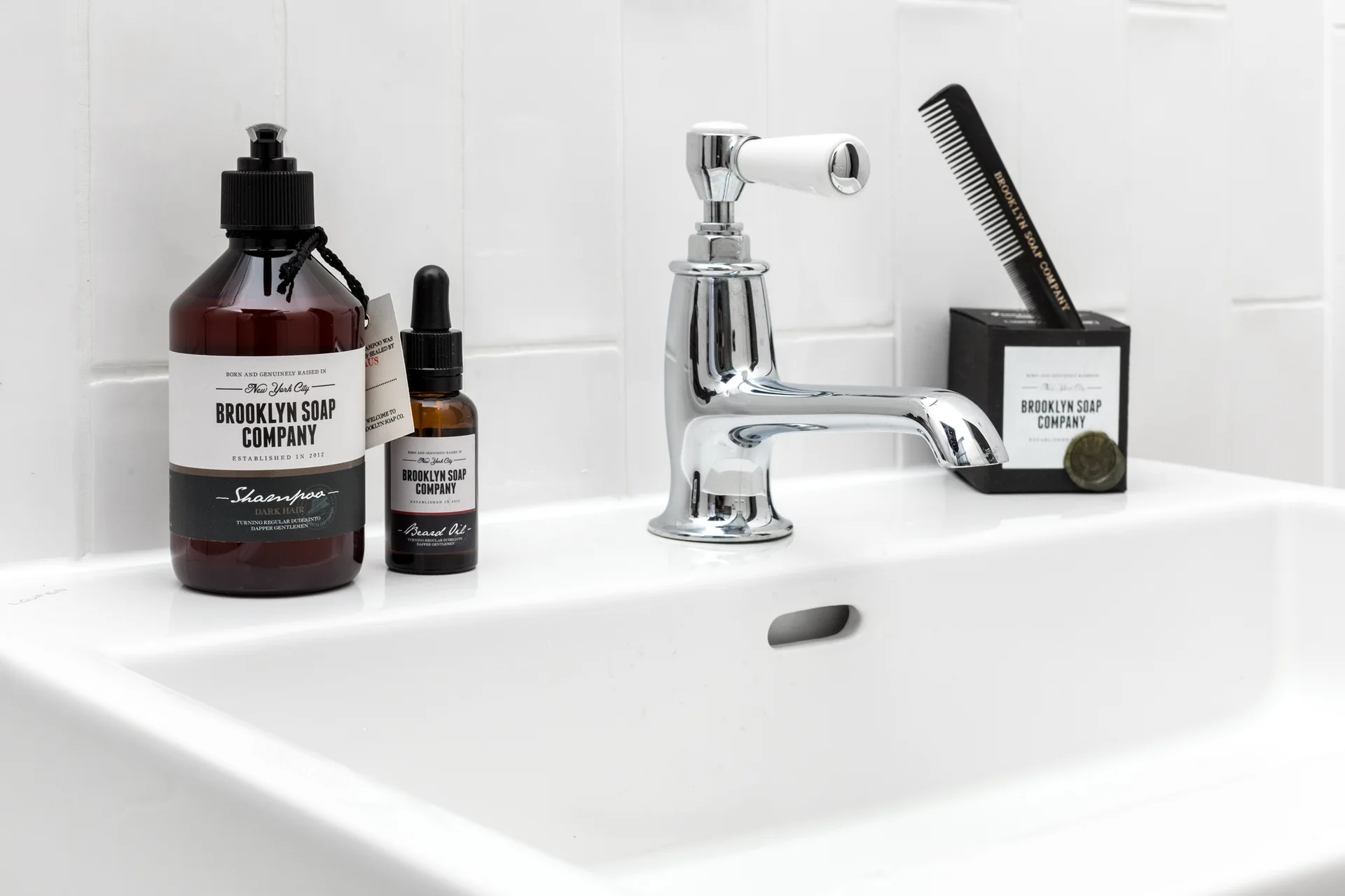 Amber glass apothecary bottles and a black comb sit on a white ceramic sink ledge below a chrome faucet and vertical tile backsplash.