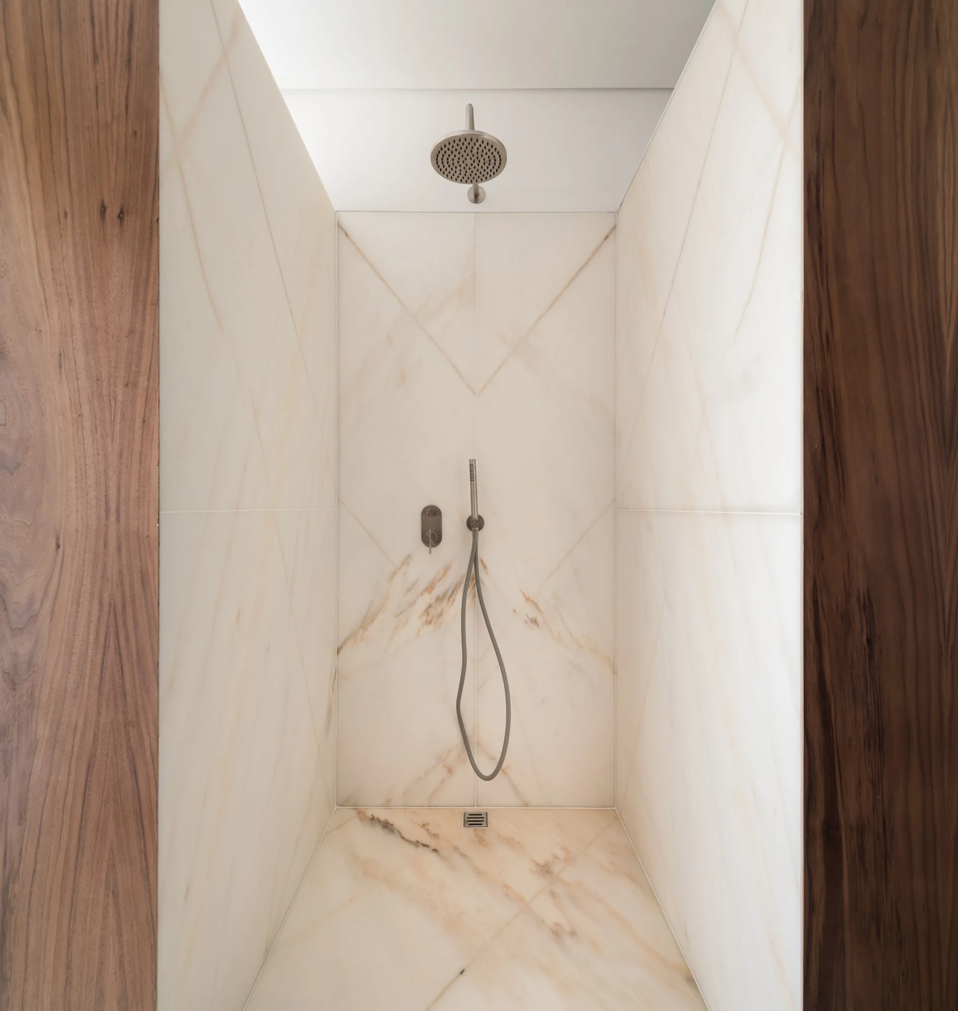A modern recessed shower enclosure lined with veined cream marble tiles and framed by vertical dark wood panels on either side.