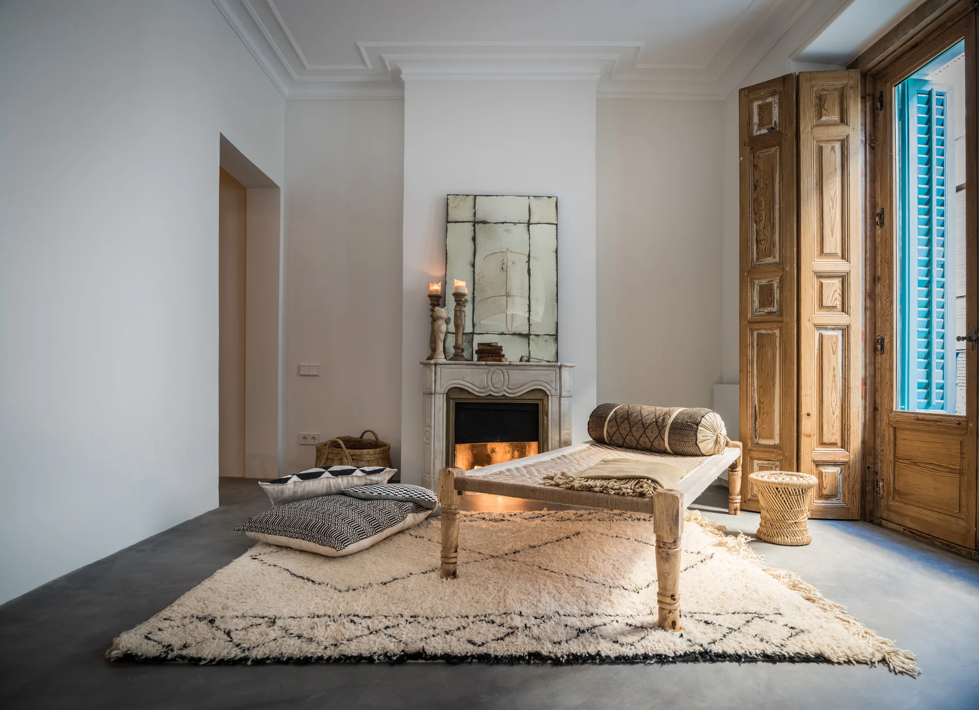 A minimalist living room featuring a stone fireplace, a woven wooden daybed, a shaggy patterned rug, and tall rustic wood folding doors.