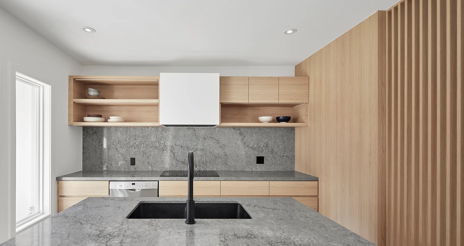 A modern kitchen featuring light wood cabinetry with open shelves, a grey stone island and matching backsplash, and a black faucet.