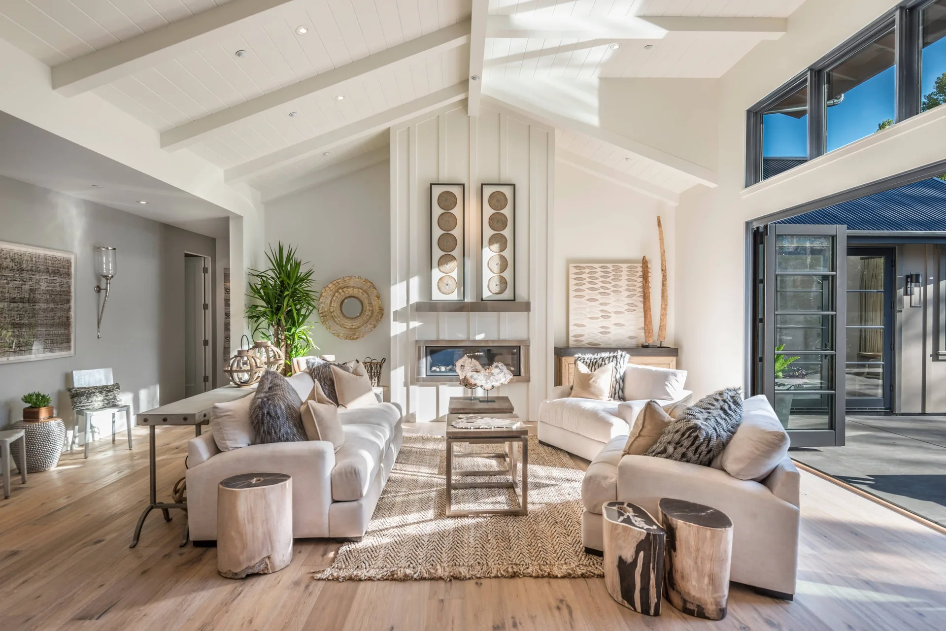A bright living room features cream sofas, natural wood stump side tables, a fireplace with vertical paneling, and a high vaulted ceiling with white beams.