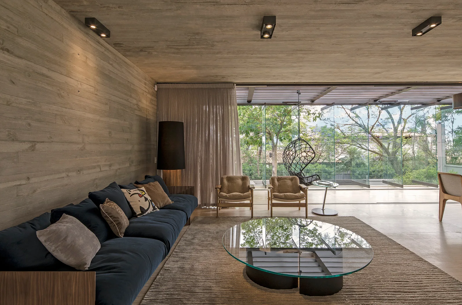 A modern living room with textured board-formed concrete walls, a large navy blue sofa, a glass coffee table, and open sliding glass doors.