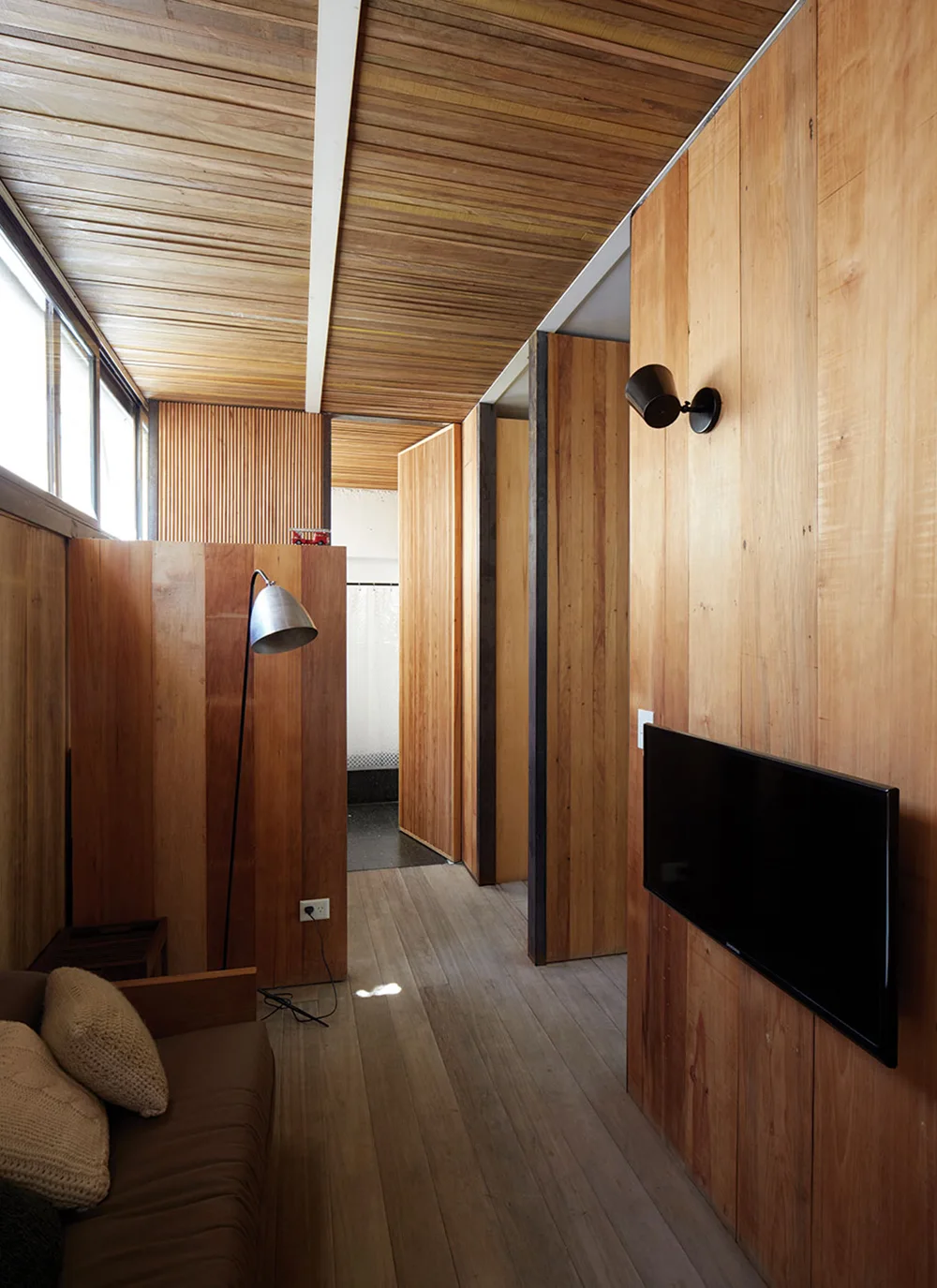 A narrow wood-paneled living room features a slatted ceiling, a brown sofa with knit pillows, and a wall-mounted television.