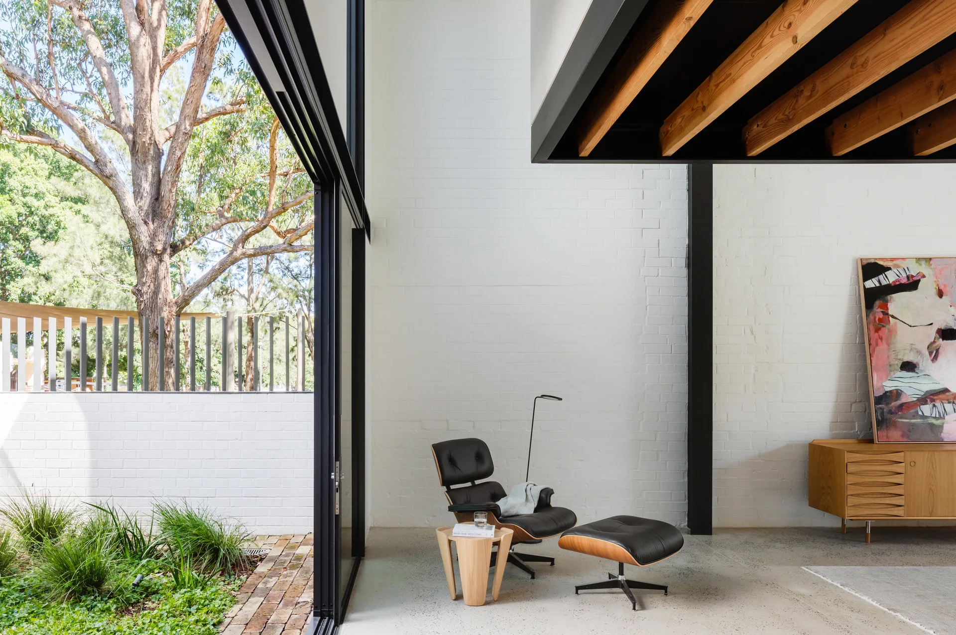 A black leather lounge chair and ottoman set on a polished concrete floor in a minimalist room with a white brick wall and sliding glass door.