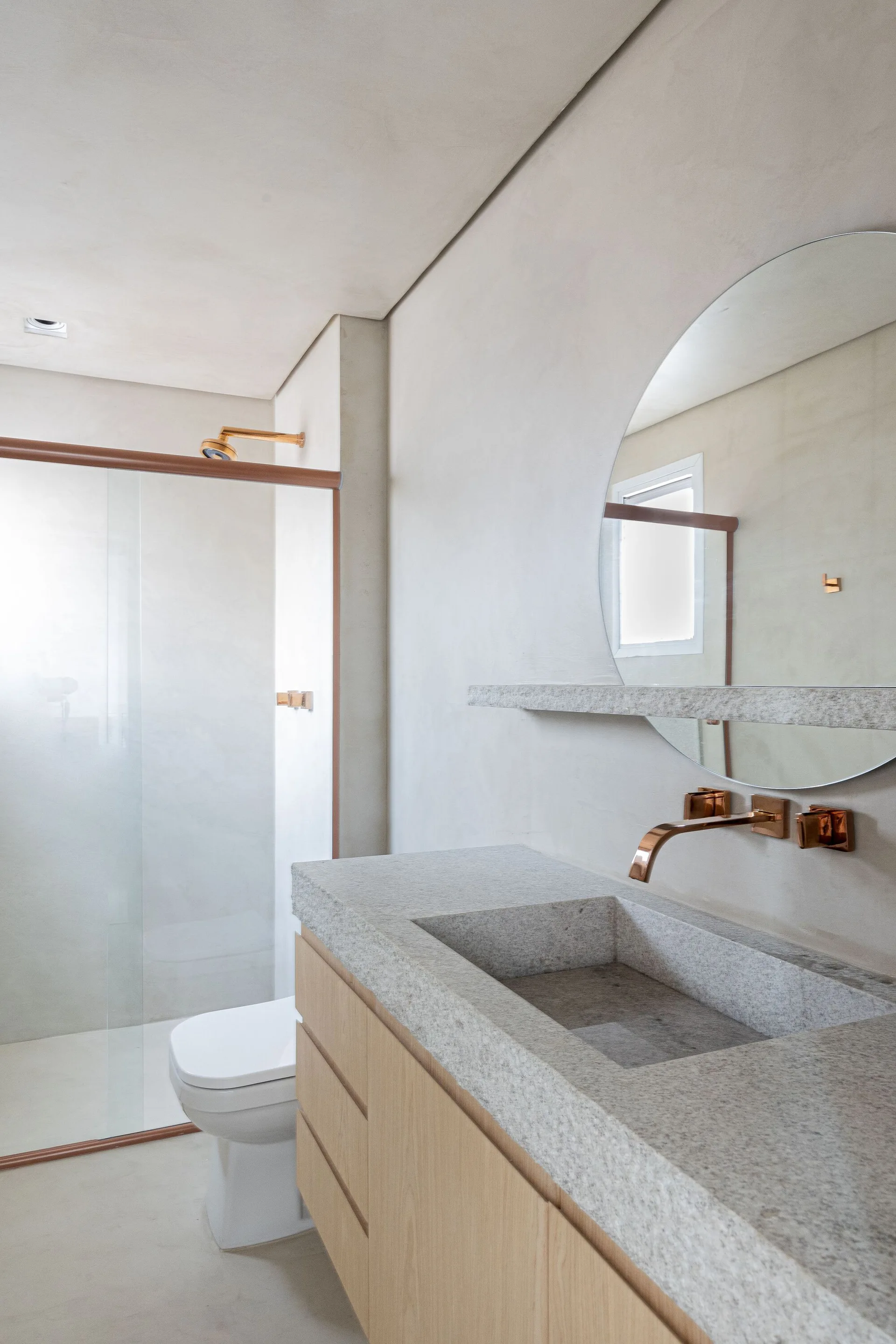 A minimalist bathroom features a light wood vanity with an integrated stone sink, copper wall-mounted faucet, and a round mirror against plaster walls.