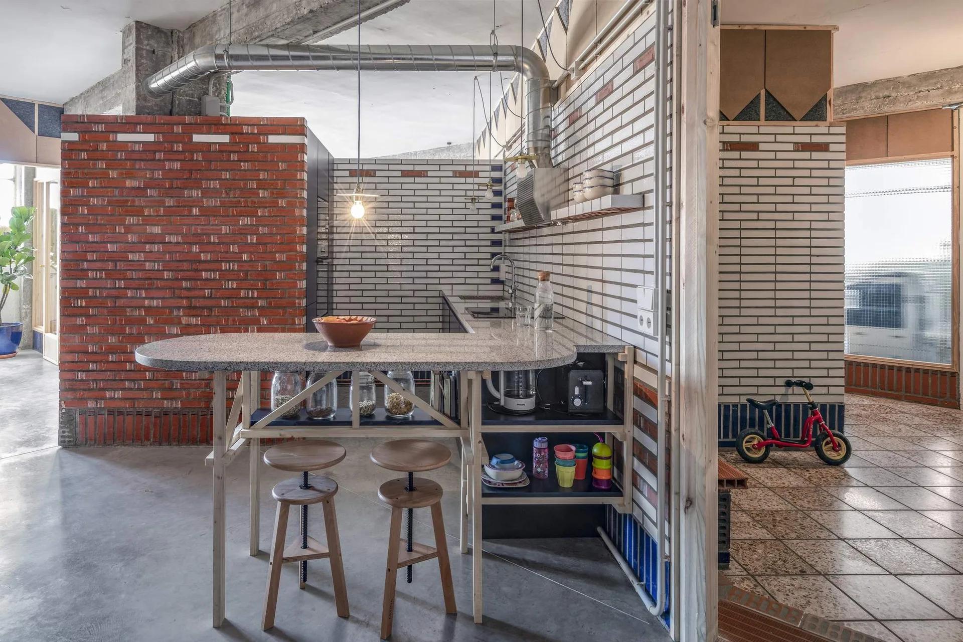 An industrial loft kitchen features a curved stone-topped island with wooden stools, set against exposed brick walls and concrete floors.