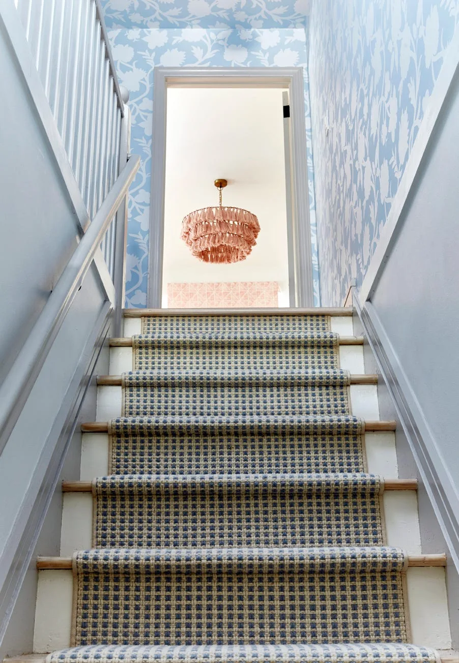 A view up a staircase featuring a blue checkered runner, floral wallpaper, and a pink tassel chandelier visible through the top doorway.