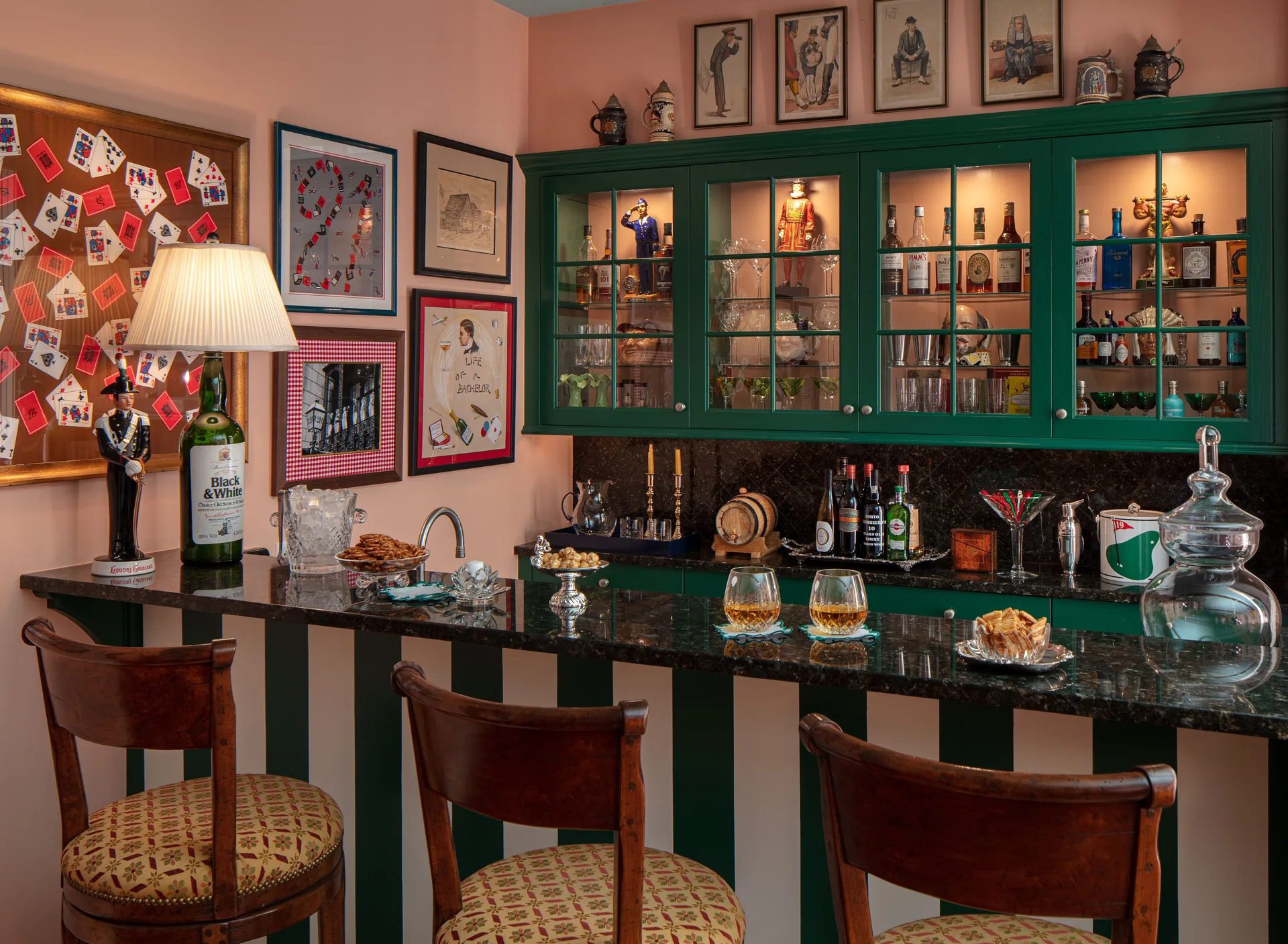 An eclectic home bar featuring green glass-front cabinets, a dark polished countertop, and a striped base paired with upholstered wooden stools.