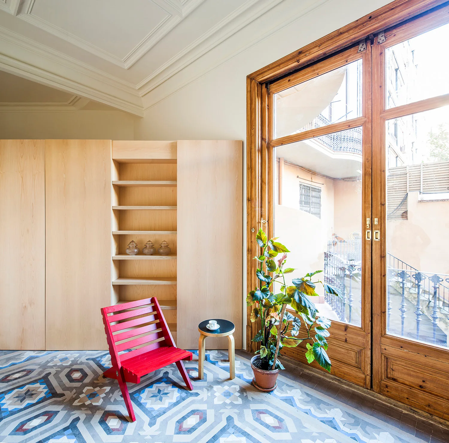 A bright room features intricate geometric floor tiles, a minimalist red slatted wood chair, and a large blonde wood storage cabinet next to glass doors.