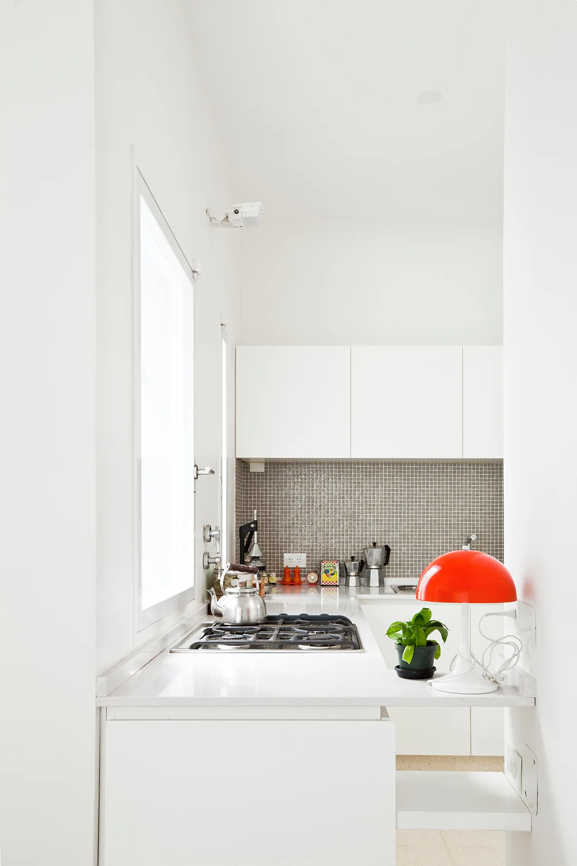 A compact white galley kitchen featuring flat-front cabinets, a grey mosaic tile backsplash, a built-in stovetop, and a red mushroom lamp.