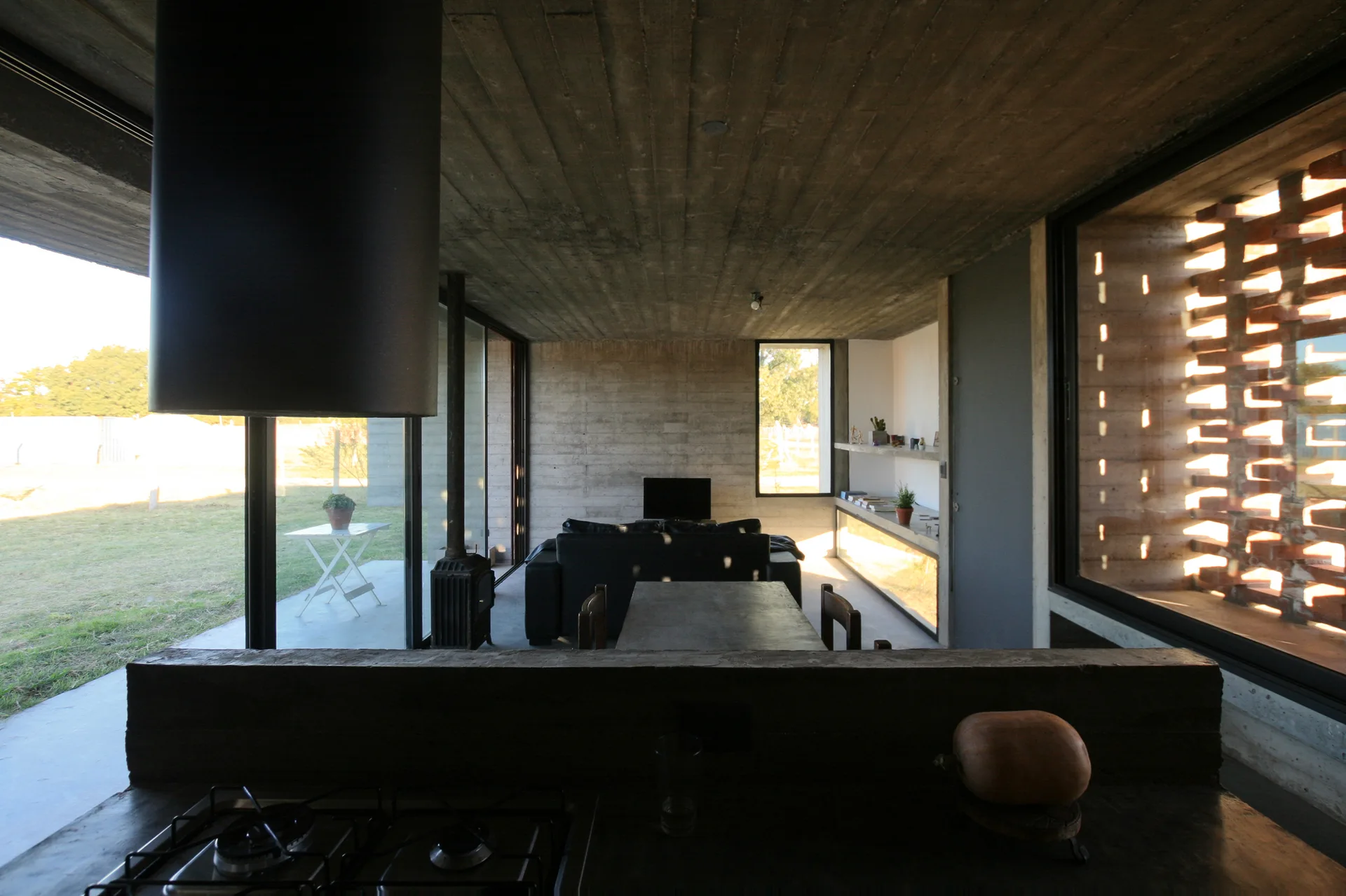 A minimalist living space featuring raw board-formed concrete walls, a dark kitchen counter, a central sofa, and a decorative brick lattice window screen.