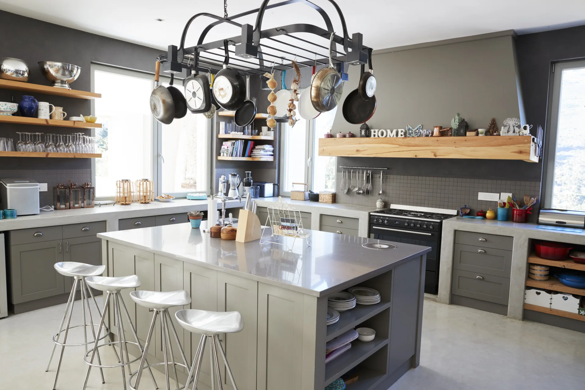 A spacious gray kitchen featuring a large central island with metal bar stools, concrete countertops, and a black hanging pot rack filled with pans.