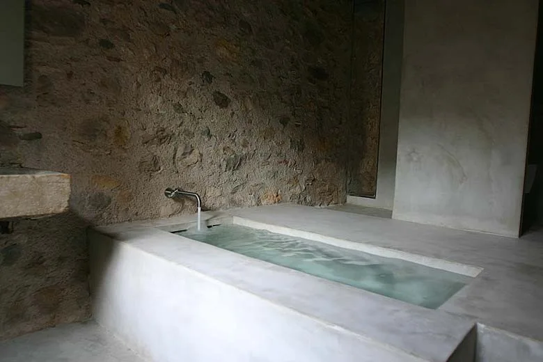 A minimalist built-in concrete bathtub is set against a rugged, natural stone wall with a single chrome faucet.