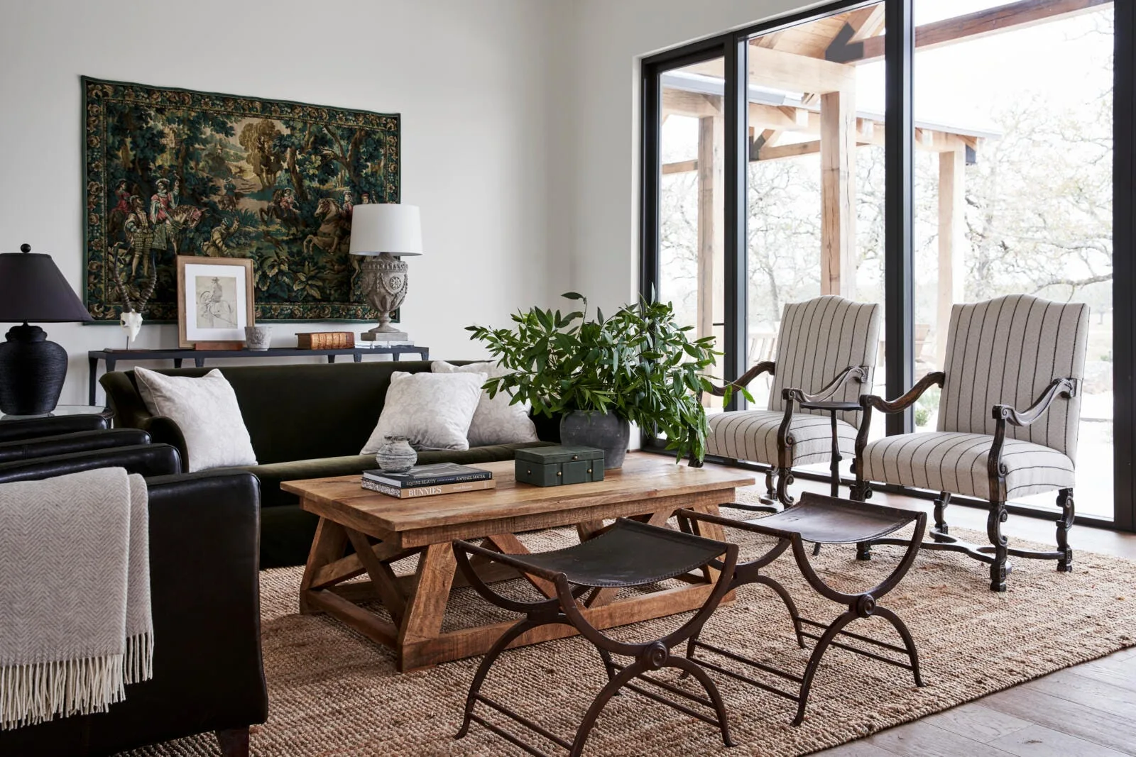 A transitional living room features a dark green velvet sofa, a wooden coffee table, striped armchairs, and a large scenic tapestry on the wall.
