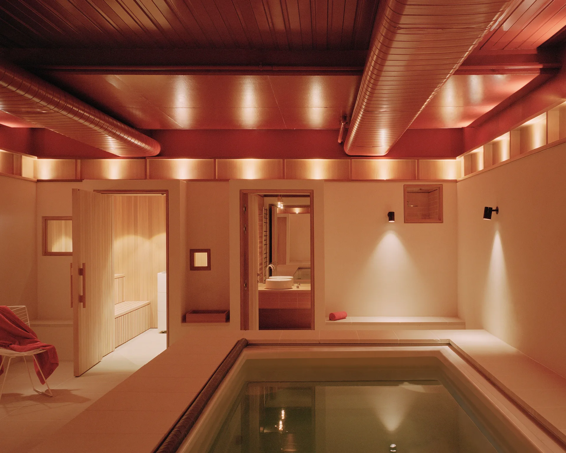 An indoor spa area with a sunken tiled pool, an open wood-lined sauna, and a warm red ceiling with exposed ductwork.