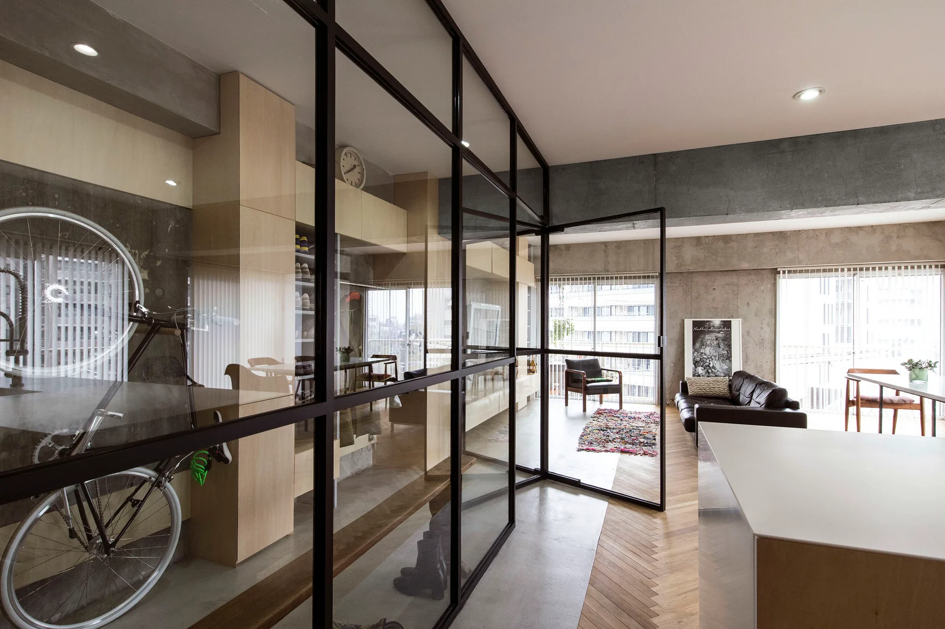 An industrial living room features a black steel-framed glass partition, exposed concrete walls, and herringbone wood flooring.